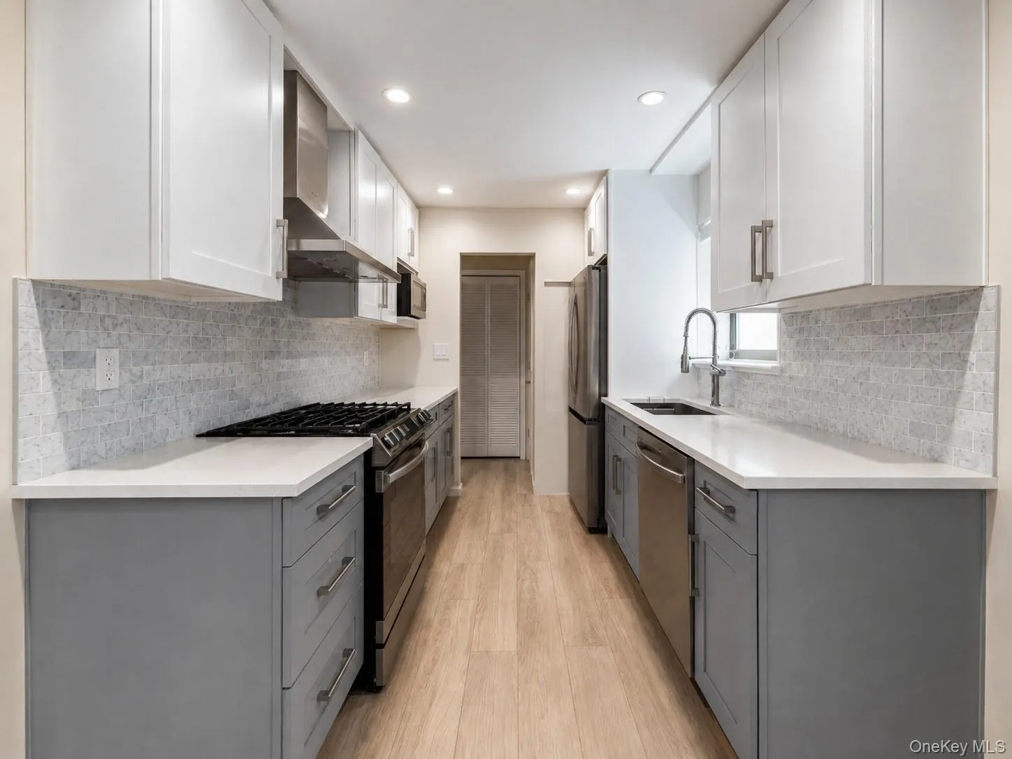 Galley kitchen featuring dual-tone cabinetry, stainless steel appliances, light countertops, a tile backsplash, and wood-finish flooring Galley kitchen featuring dual-tone cabinetry, stainless steel appliances, light countertops, a tile backsplash, and wood-finish flooring