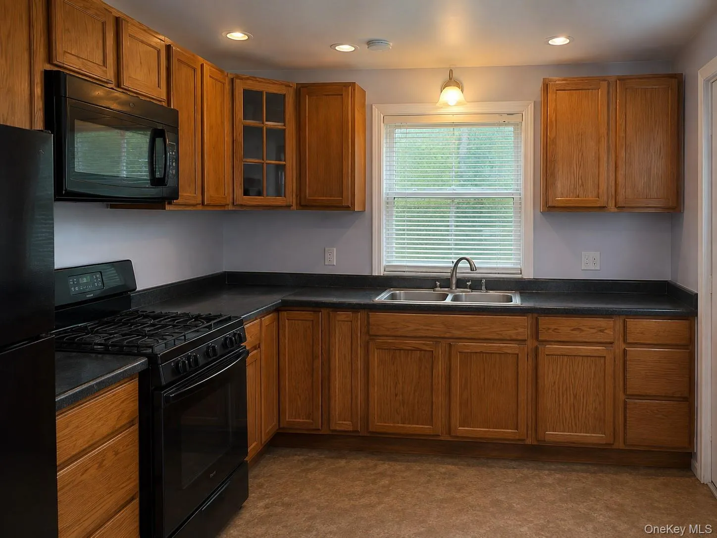 Spacious kitchen featuring warm wood cabinetry with glass-front upper doors, black gas range with ***Photos have been digitally staged to remove clutter***matching black microwave, dark countertops, stainless steel sink with single-handle faucet, recessed ceiling lighting, and a large window with plantation shutters providing natural light Spacious kitchen featuring warm wood cabinetry with glass-front upper doors, black gas range with ***Photos have been digitally staged to remove clutter***matching black microwave, dark countertops, stainless steel sink with single-handle faucet, recessed ceiling lighting, and a large window with plantation shutters providing natural light
