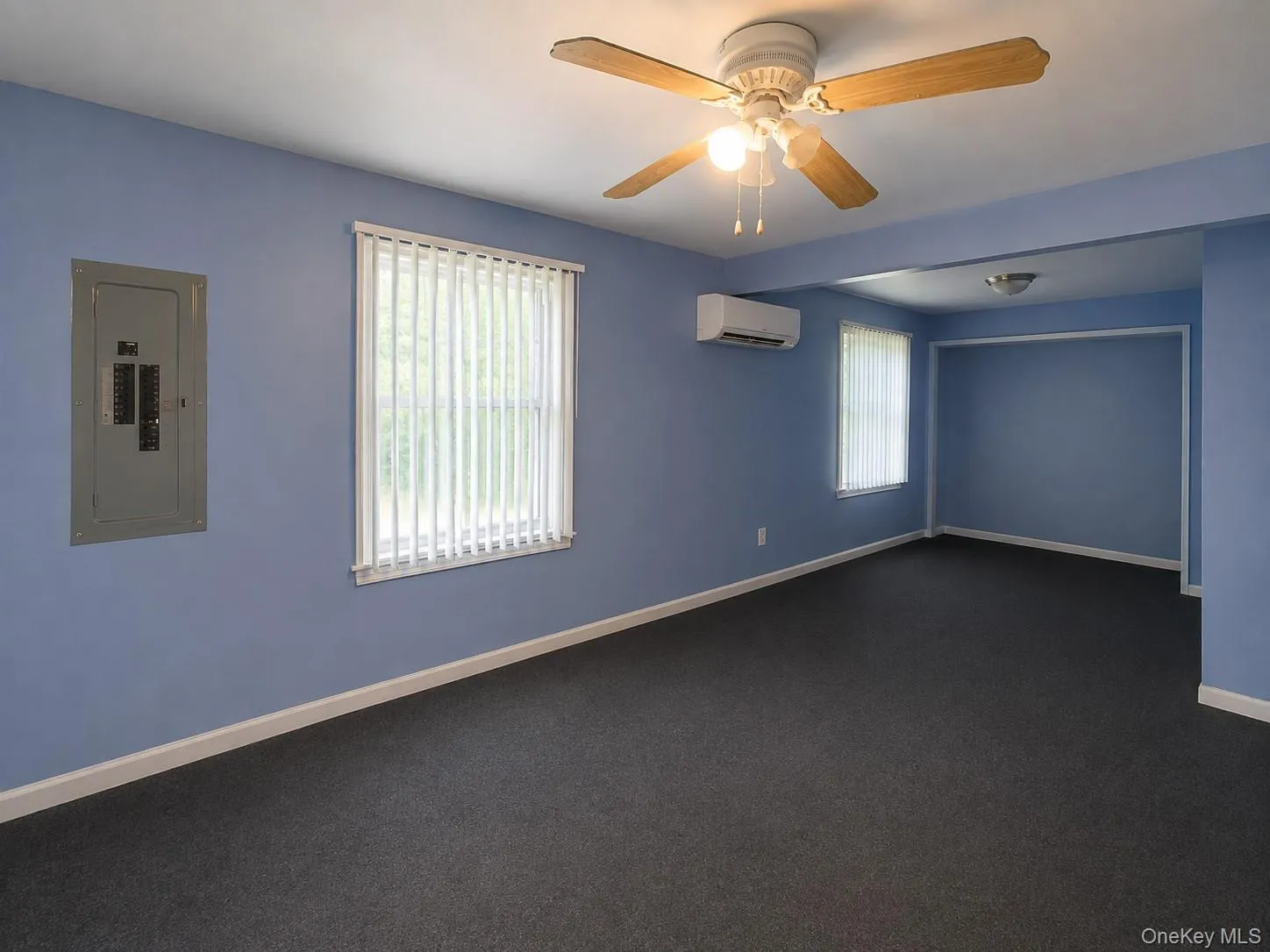 Spacious room featuring perimeter windows with vertical blinds, neutral wall-to-wall carpeting, ceiling fan with integrated lighting, wall-mounted air conditioning unit, and fresh blue-painted walls with white trim***Photos have been digitally staged to remove clutter*** Spacious room featuring perimeter windows with vertical blinds, neutral wall-to-wall carpeting, ceiling fan with integrated lighting, wall-mounted air conditioning unit, and fresh blue-painted walls with white trim***Photos have been digitally staged to remove clutter***