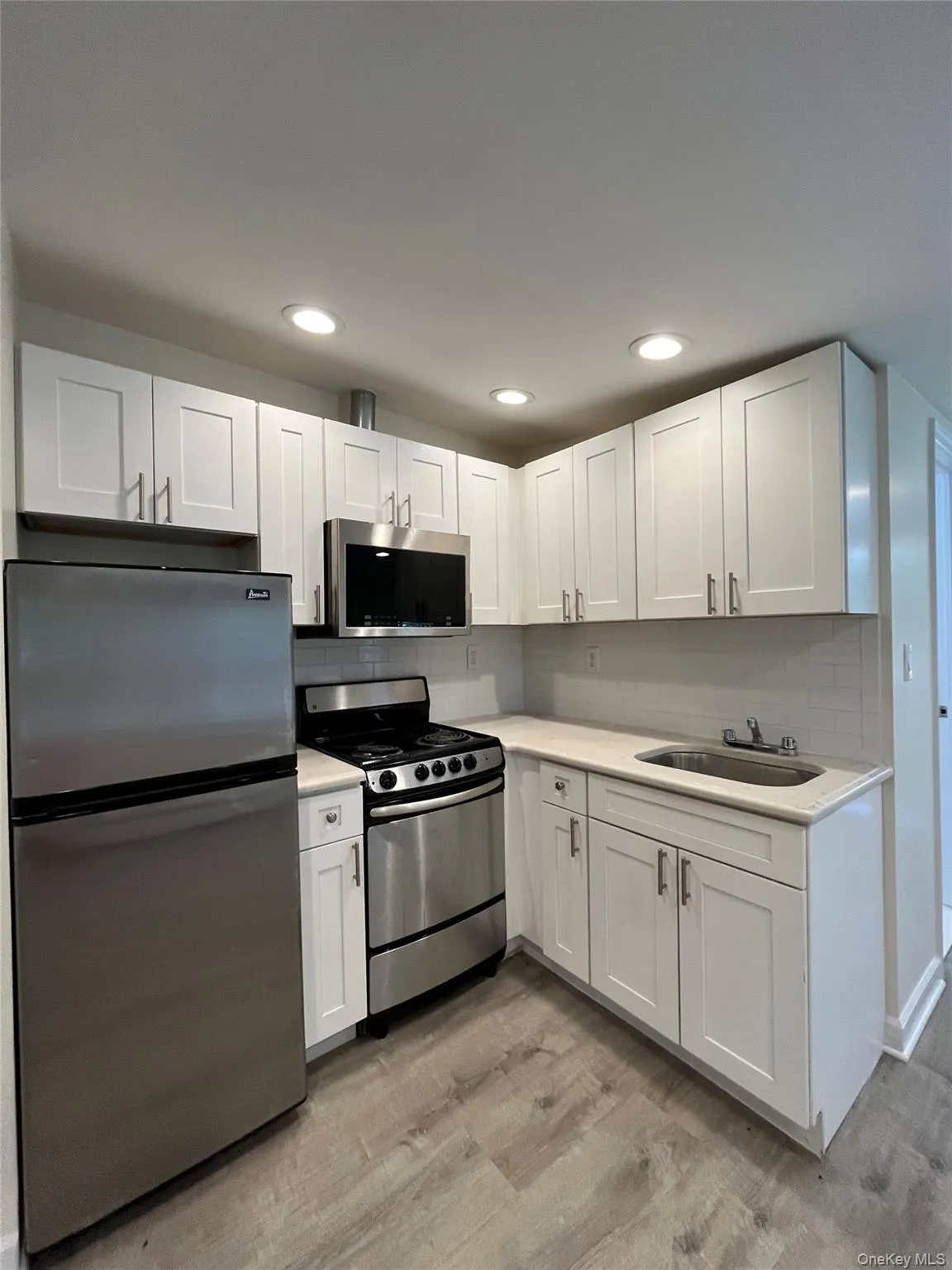 Contemporary kitchen featuring stainless steel appliances, white shaker-style cabinetry, light-toned countertops, a subway tile backsplash, and wood-finish flooring Contemporary kitchen featuring stainless steel appliances, white shaker-style cabinetry, light-toned countertops, a subway tile backsplash, and wood-finish flooring