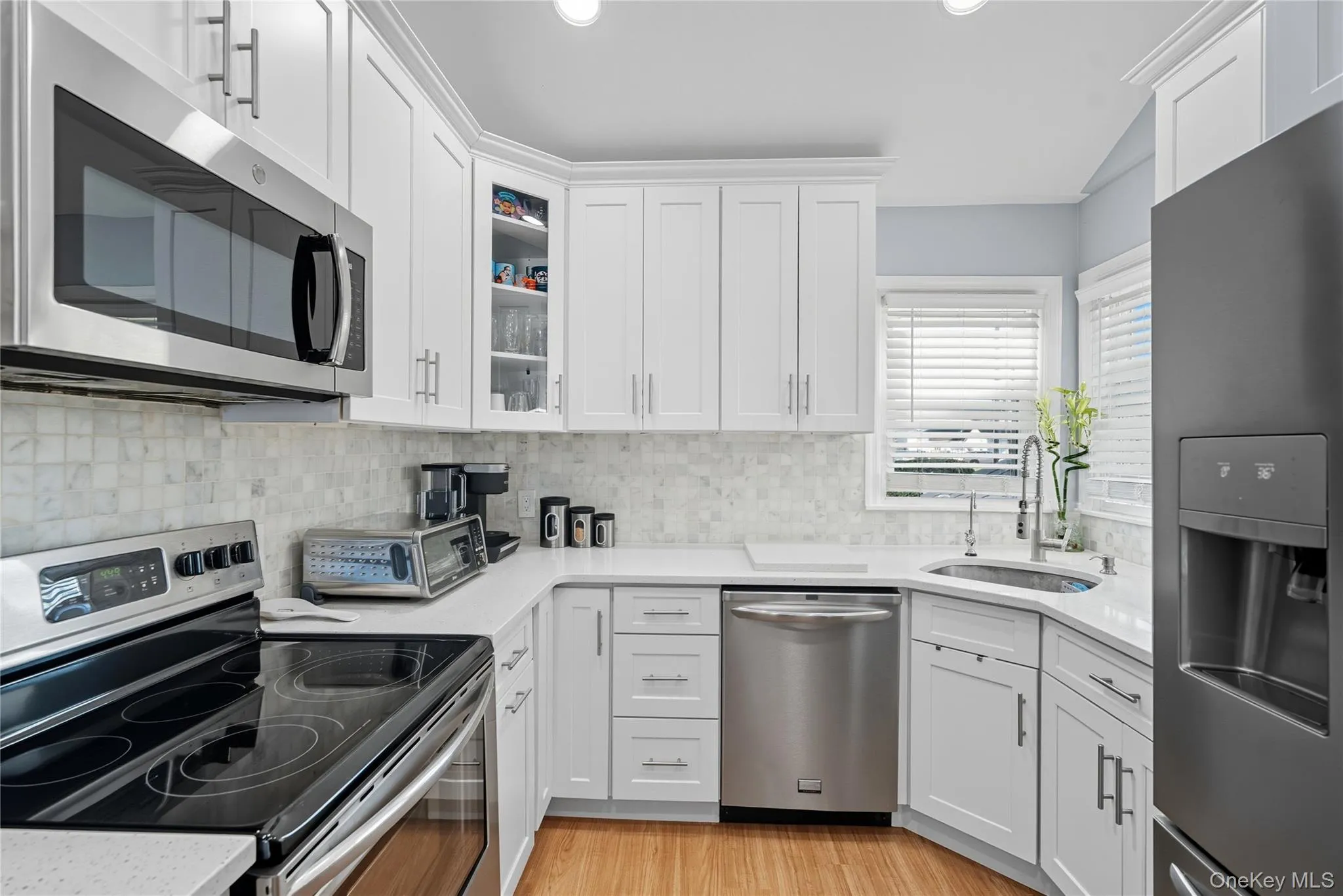 Kitchen with appliances with stainless steel finishes, white cabinetry, backsplash, glass insert cabinets, and recessed lighting Kitchen with appliances with stainless steel finishes, white cabinetry, backsplash, glass insert cabinets, and recessed lighting