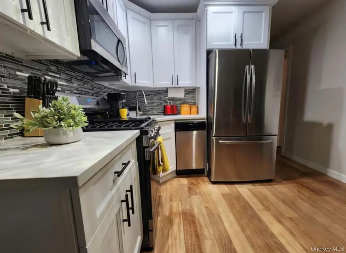 White shaker-style cabinetry with matte black hardware, stainless steel appliances, a multi-tone glass tile backsplash, light-toned countertops, and wood-finish flooring White shaker-style cabinetry with matte black hardware, stainless steel appliances, a multi-tone glass tile backsplash, light-toned countertops, and wood-finish flooring