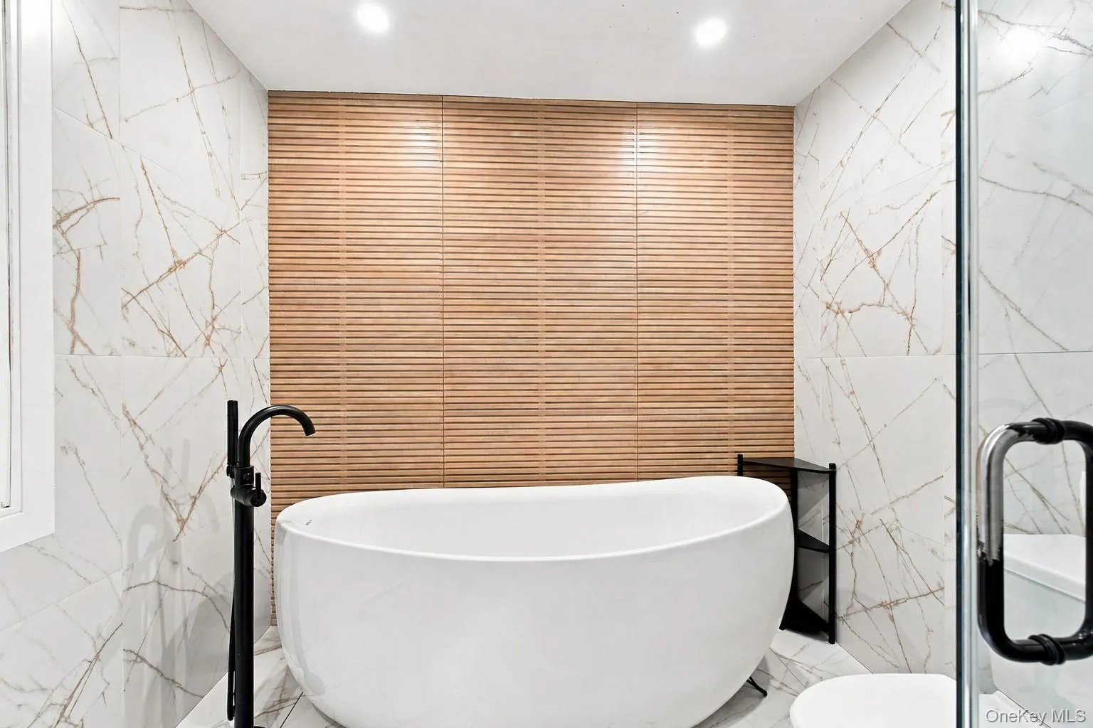 Freestanding soaking tub with a floor-mounted matte black faucet, wall-mounted wood-finish tile, and large format white tile with gold veining Freestanding soaking tub with a floor-mounted matte black faucet, wall-mounted wood-finish tile, and large format white tile with gold veining