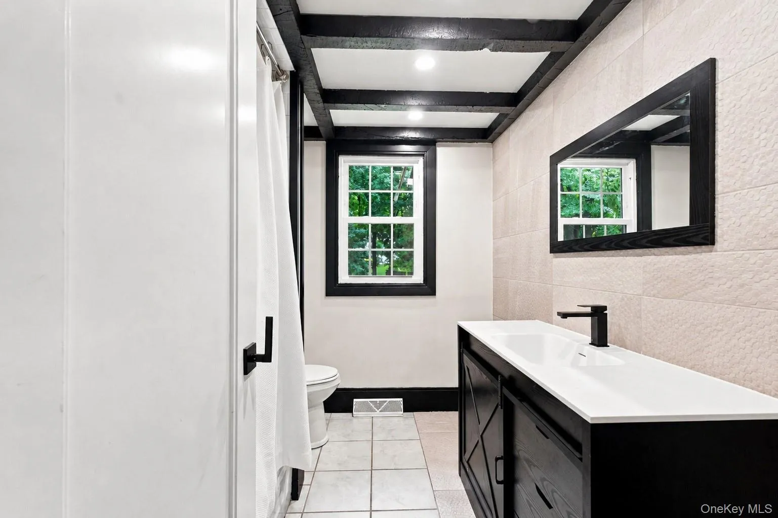 Bathroom featuring exposed ceiling beams, horizontal-set wall tile, a window with dark trim, a dark vanity with an integrated sink, and matte black fixtures Bathroom featuring exposed ceiling beams, horizontal-set wall tile, a window with dark trim, a dark vanity with an integrated sink, and matte black fixtures