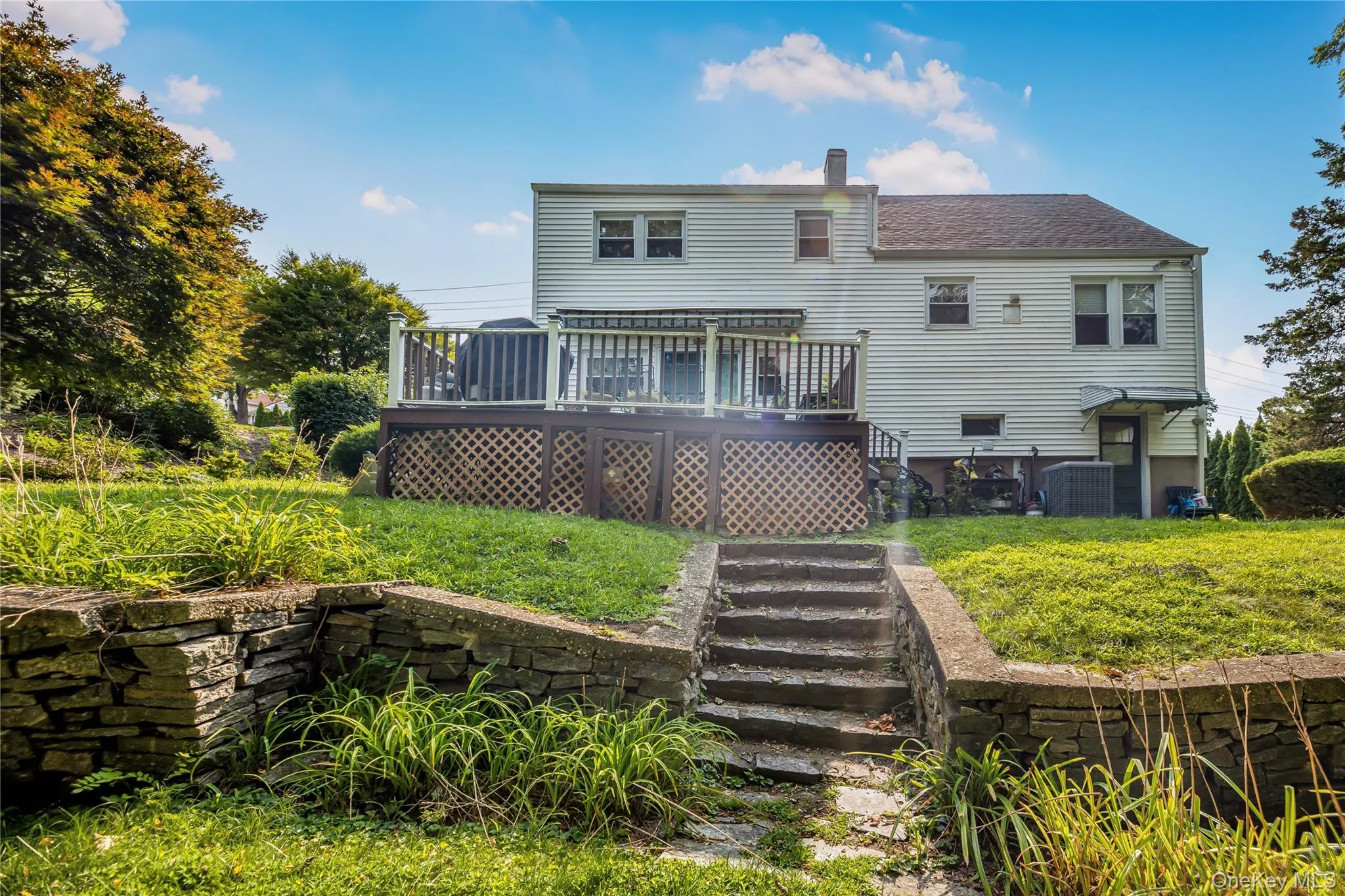 Rear exterior featuring a multi-level wood deck with lattice skirting, white siding, and a stone retaining wall Rear exterior featuring a multi-level wood deck with lattice skirting, white siding, and a stone retaining wall