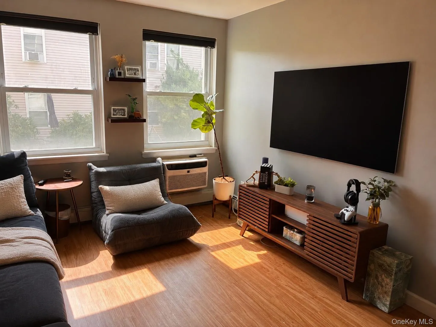 Living area featuring laminate flooring, two large windows with shades, wall mounted AC unit and a wall-mounted entertainment unit Living area featuring laminate flooring, two large windows with shades, wall mounted AC unit and a wall-mounted entertainment unit