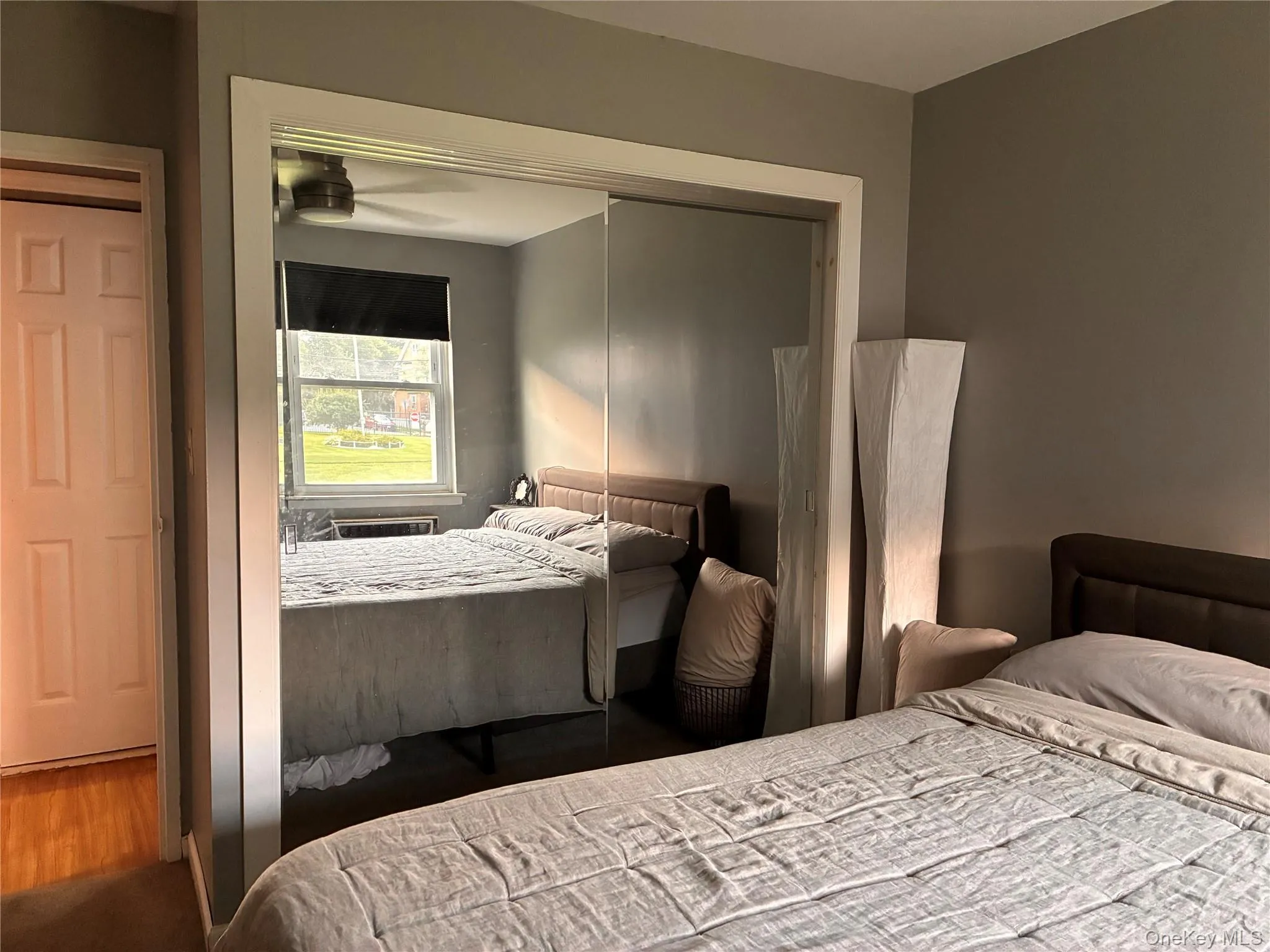 Bedroom with integrated ceiling fan, single window with shade, wall mounted AC unit, double closet with mirrored sliding doors, and neutral wall paint Bedroom with integrated ceiling fan, single window with shade, wall mounted AC unit, double closet with mirrored sliding doors, and neutral wall paint