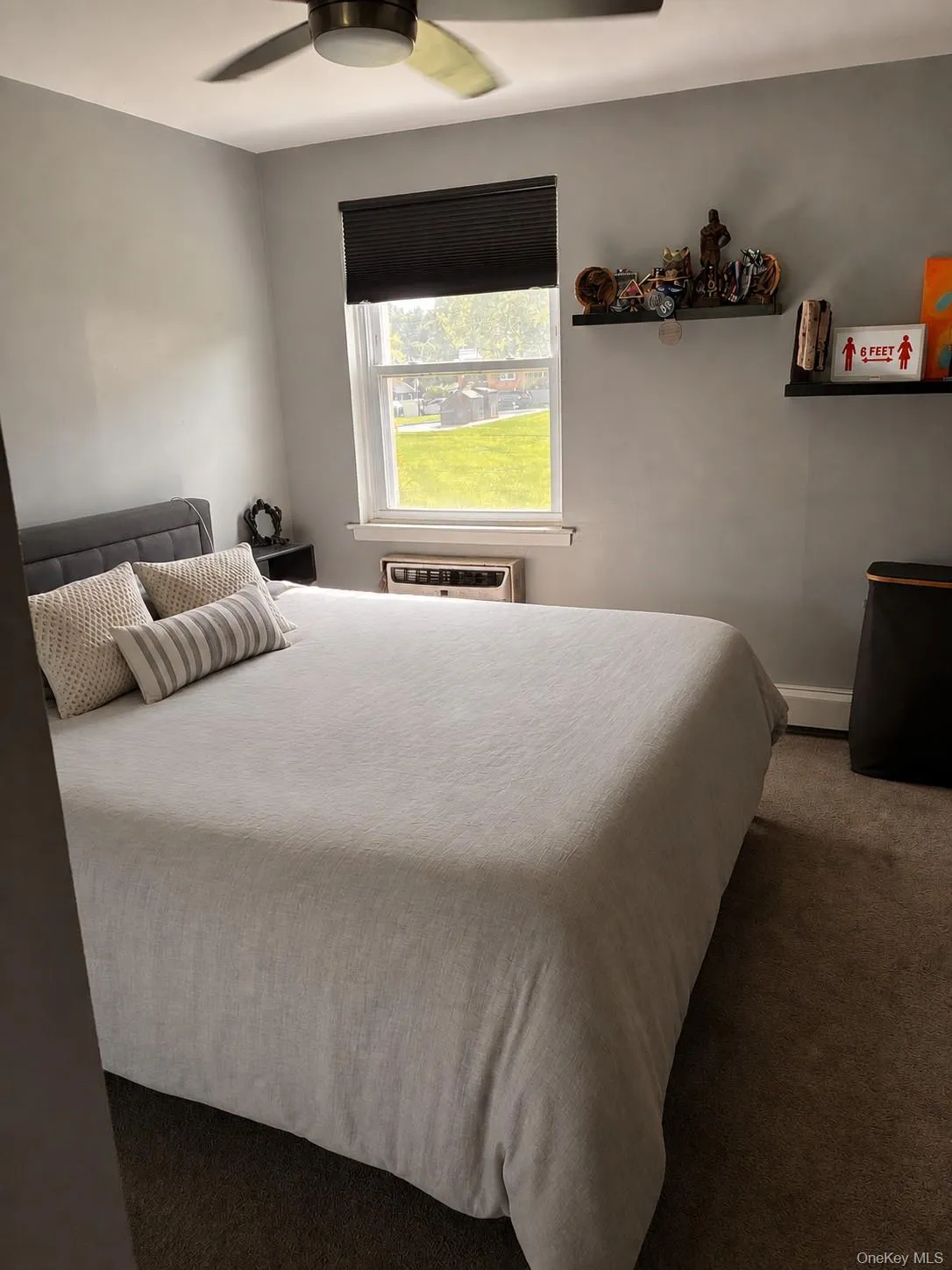 Bedroom with integrated ceiling fan, single window with shade, wall mounted AC unit, shelving, and neutral wall paint Bedroom with integrated ceiling fan, single window with shade, wall mounted AC unit, shelving, and neutral wall paint