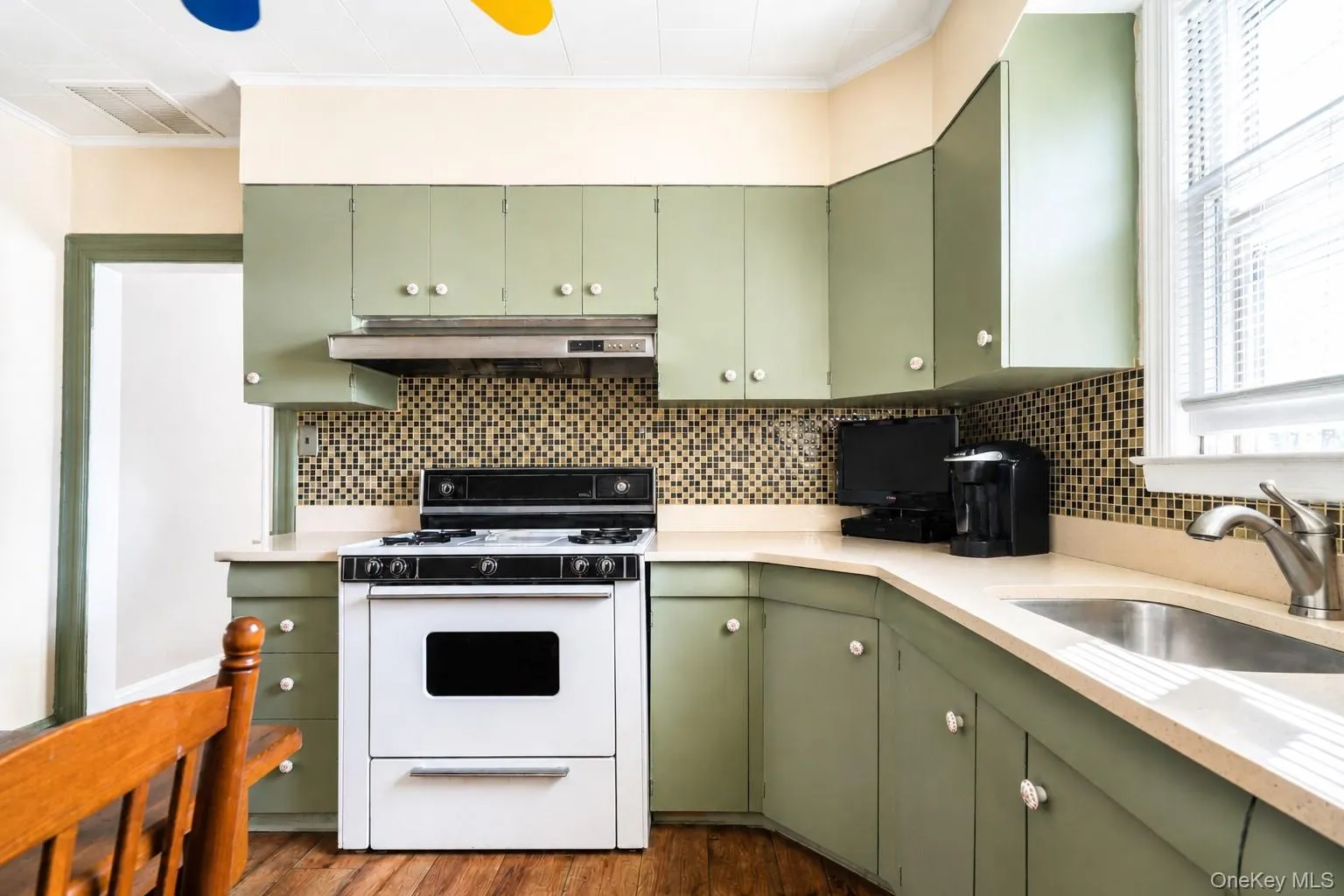 Charming kitchen featuring sage-green cabinetry with brushed nickel hardware, white subway tile backsplash with geometric accent pattern, white enamel range with gas cooktop, stainless steel sink with chrome faucet, and cream-colored countertops Charming kitchen featuring sage-green cabinetry with brushed nickel hardware, white subway tile backsplash with geometric accent pattern, white enamel range with gas cooktop, stainless steel sink with chrome faucet, and cream-colored countertops