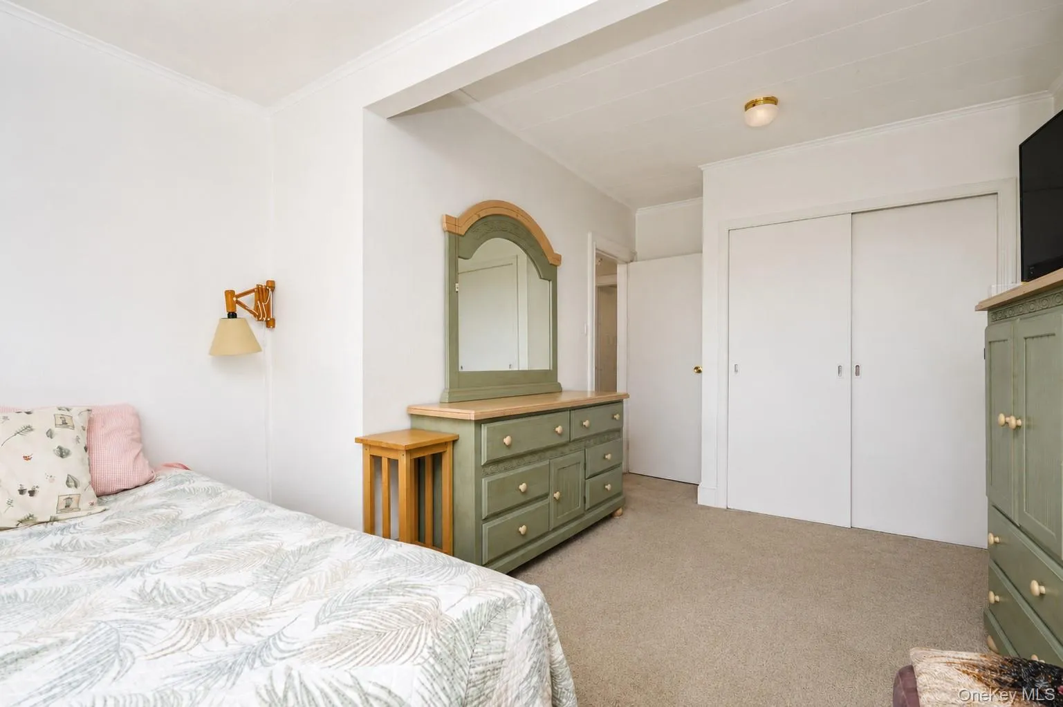 Spacious bedroom featuring neutral carpeting, wood-finish dresser with arched mirror, warm brass wall sconce, and direct access to multiple closets with white panel doors Spacious bedroom featuring neutral carpeting, wood-finish dresser with arched mirror, warm brass wall sconce, and direct access to multiple closets with white panel doors