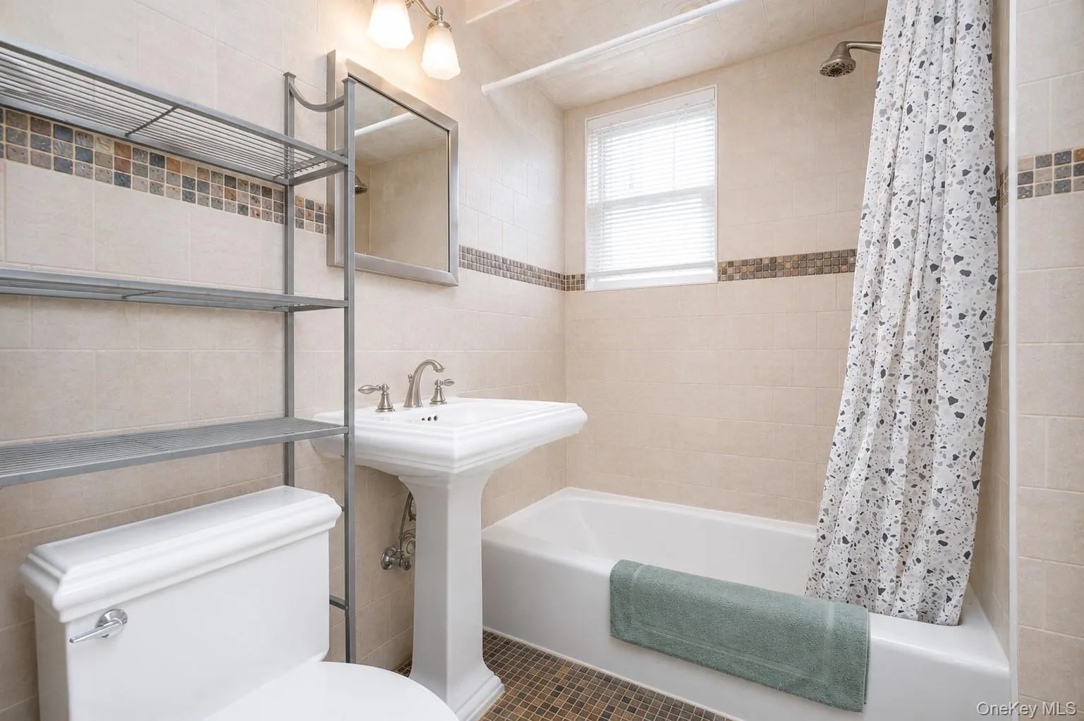 Bright bathroom featuring a pedestal sink with chrome faucet, white subway tile walls with decorative tile border, ceramic tile flooring, toilet with dual-flush mechanism, bathtub with shower enclosure and patterned shower curtain, wall-mounted mirror with chrome frame, and open shelving with towel storage Bright bathroom featuring a pedestal sink with chrome faucet, white subway tile walls with decorative tile border, ceramic tile flooring, toilet with dual-flush mechanism, bathtub with shower enclosure and patterned shower curtain, wall-mounted mirror with chrome frame, and open shelving with towel storage