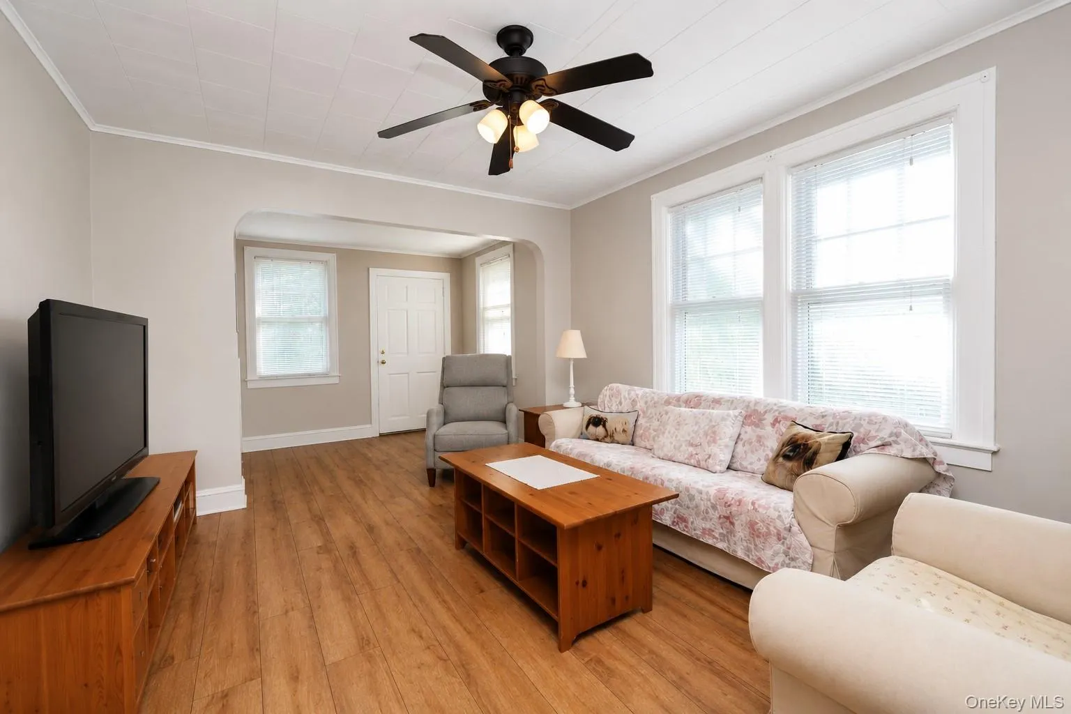 Wood-finish flooring throughout, ceiling fan with integrated lighting, multiple windows, and an arched doorway Wood-finish flooring throughout, ceiling fan with integrated lighting, multiple windows, and an arched doorway