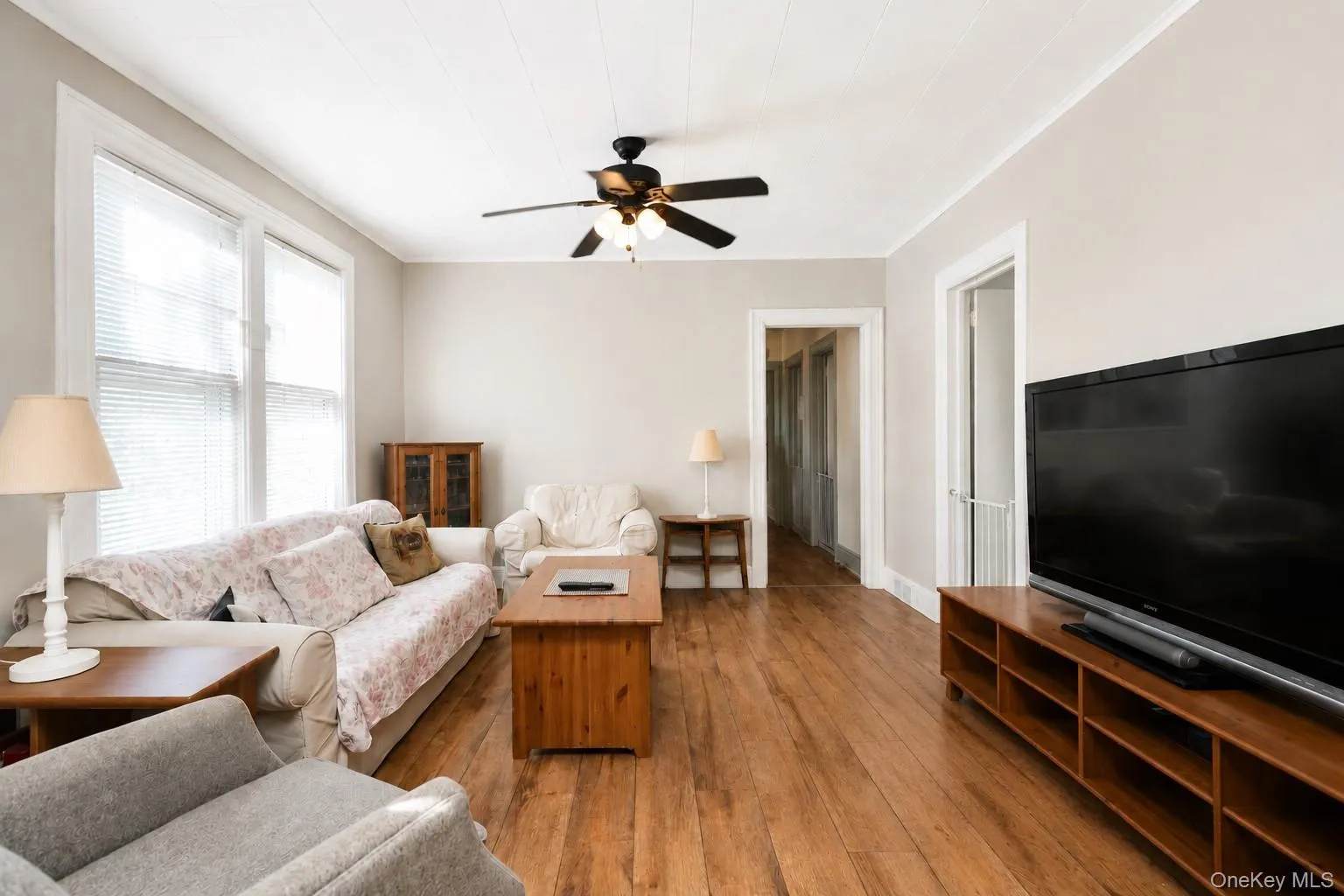 Spacious living room showcasing warm wood-finish flooring, ceiling fan with integrated lighting, large flat-screen television mounted on built-in cabinetry with open shelving, multiple windows with natural light, and comfortable seating arrangements throughout the room Spacious living room showcasing warm wood-finish flooring, ceiling fan with integrated lighting, large flat-screen television mounted on built-in cabinetry with open shelving, multiple windows with natural light, and comfortable seating arrangements throughout the room