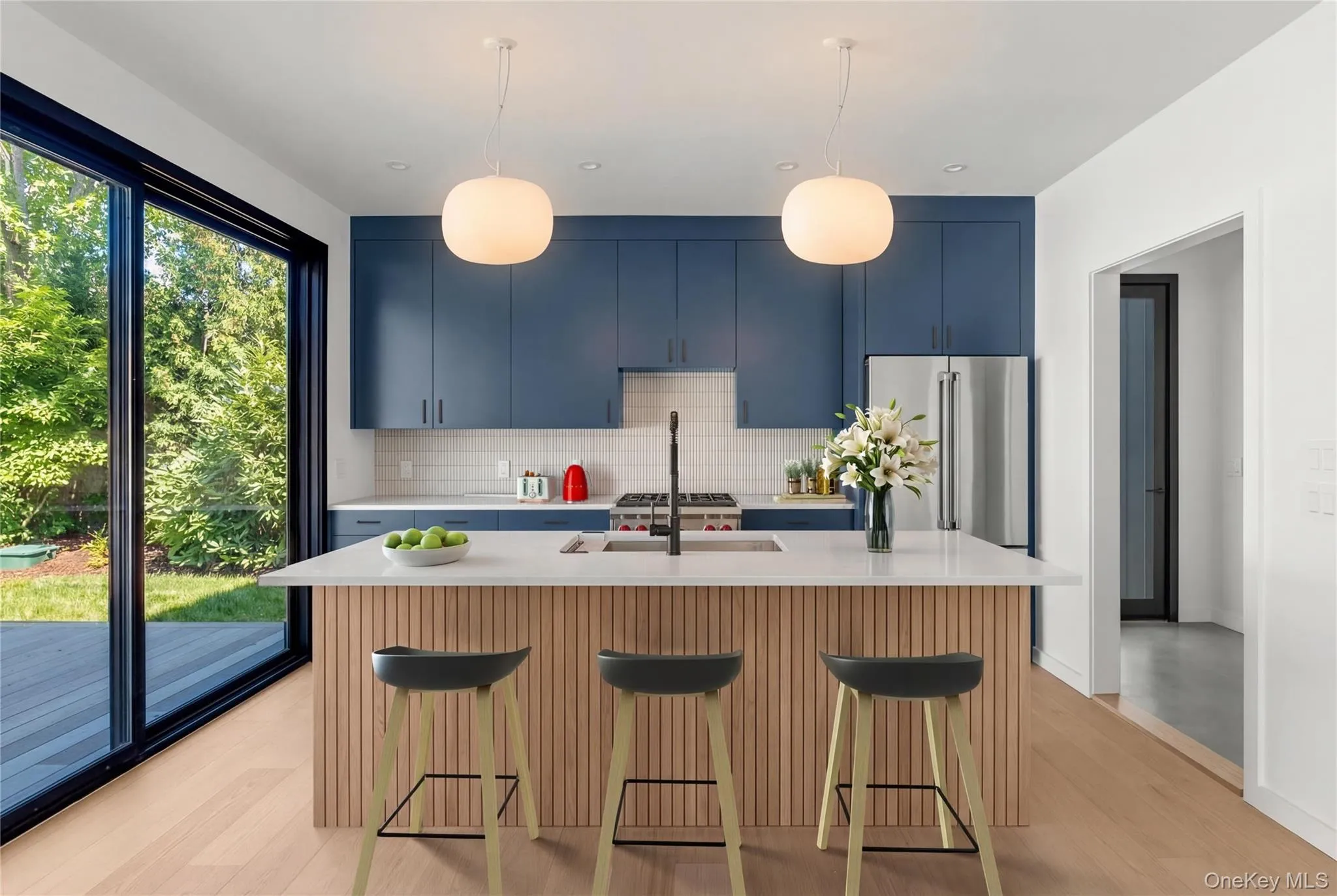 Modern kitchen featuring blue cabinetry, light wood-finish flooring, a central island with a white countertop, stainless steel appliances, and large sliding glass doors opening to the exterior Modern kitchen featuring blue cabinetry, light wood-finish flooring, a central island with a white countertop, stainless steel appliances, and large sliding glass doors opening to the exterior