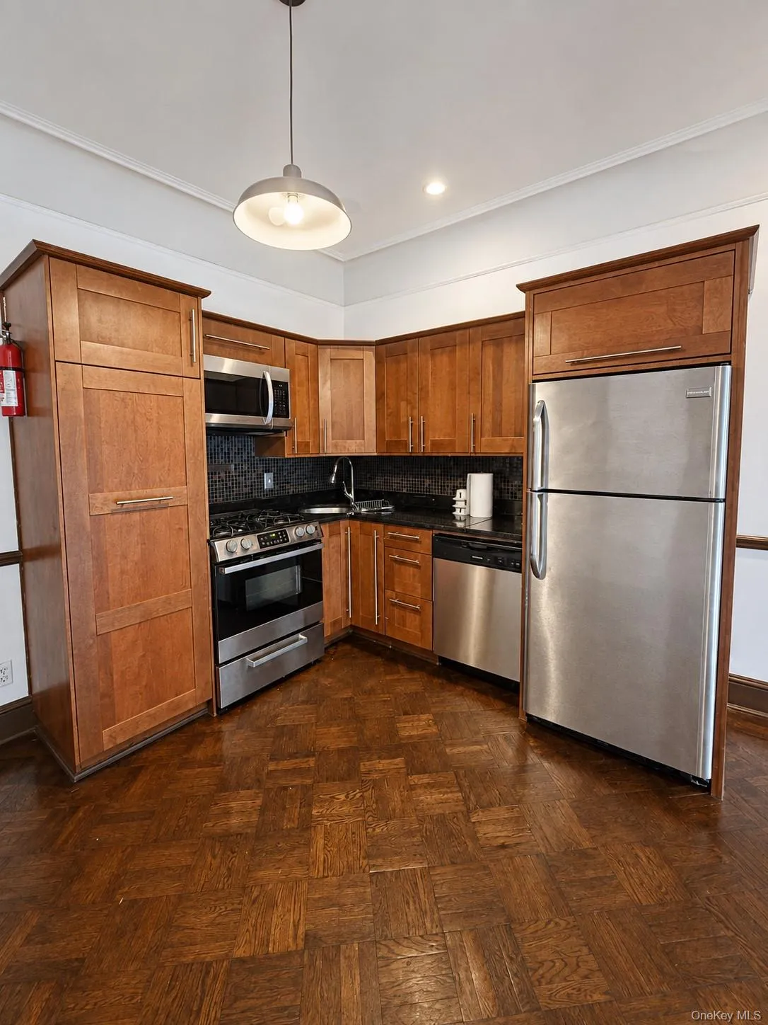 Efficient galley kitchen featuring warm wood cabinetry, stainless steel appliances including gas range and refrigerator, black countertops, pendant lighting, and rich wood-finish flooring throughout Efficient galley kitchen featuring warm wood cabinetry, stainless steel appliances including gas range and refrigerator, black countertops, pendant lighting, and rich wood-finish flooring throughout