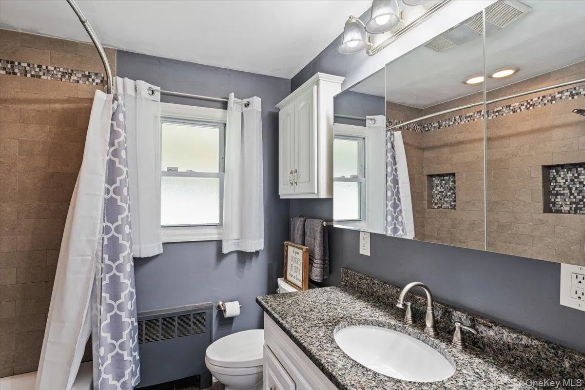 Granite countertop with oval sink and brushed nickel faucet, large mirrored medicine cabinet with overhead lighting, tiled shower surround with mosaic accent, built-in shower niches, and white wall cabinet Granite countertop with oval sink and brushed nickel faucet, large mirrored medicine cabinet with overhead lighting, tiled shower surround with mosaic accent, built-in shower niches, and white wall cabinet