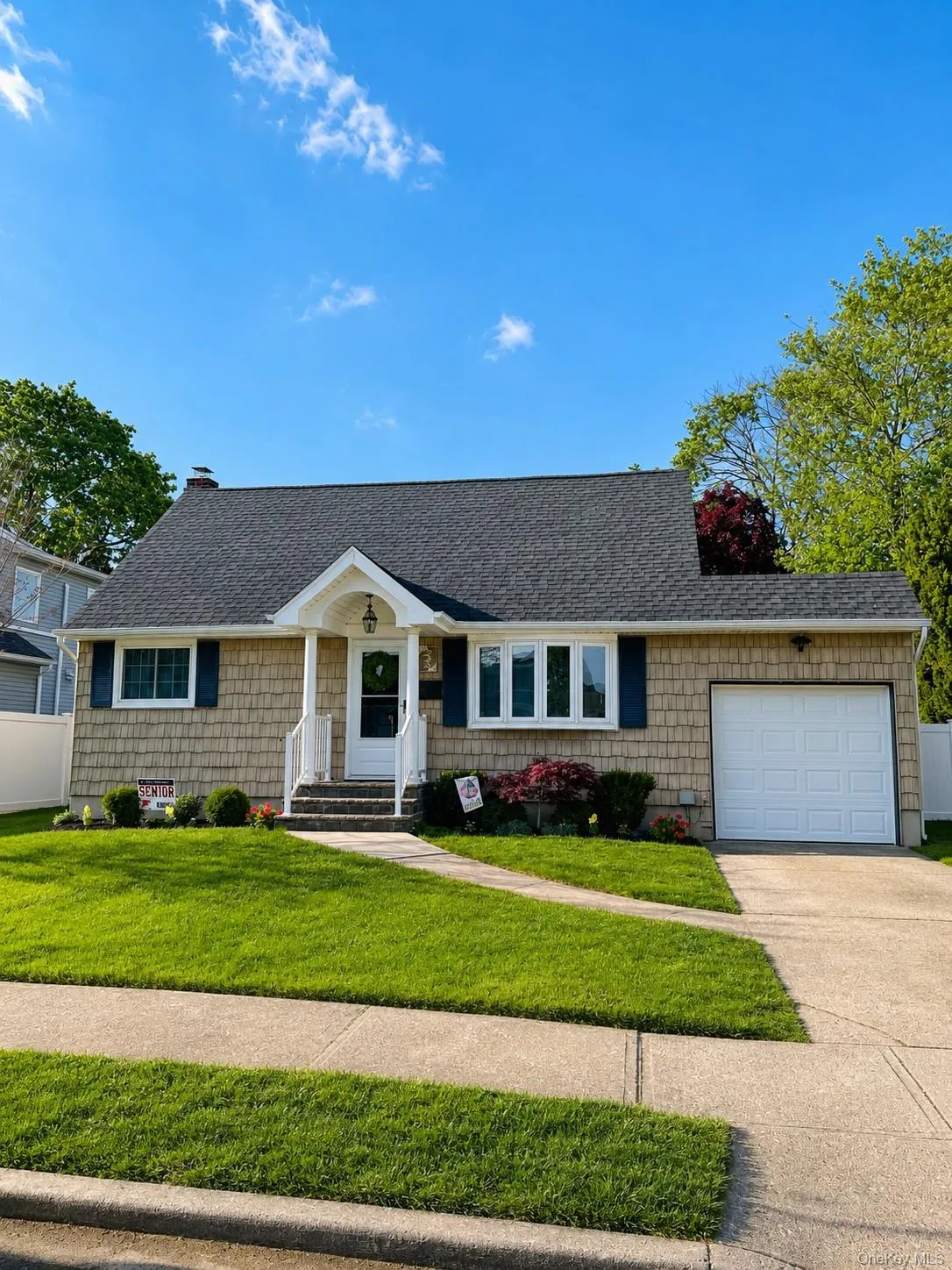 Shingle siding facade with dark blue shutters, covered entry with white railing, attached garage, paved driveway, and established front lawn Shingle siding facade with dark blue shutters, covered entry with white railing, attached garage, paved driveway, and established front lawn