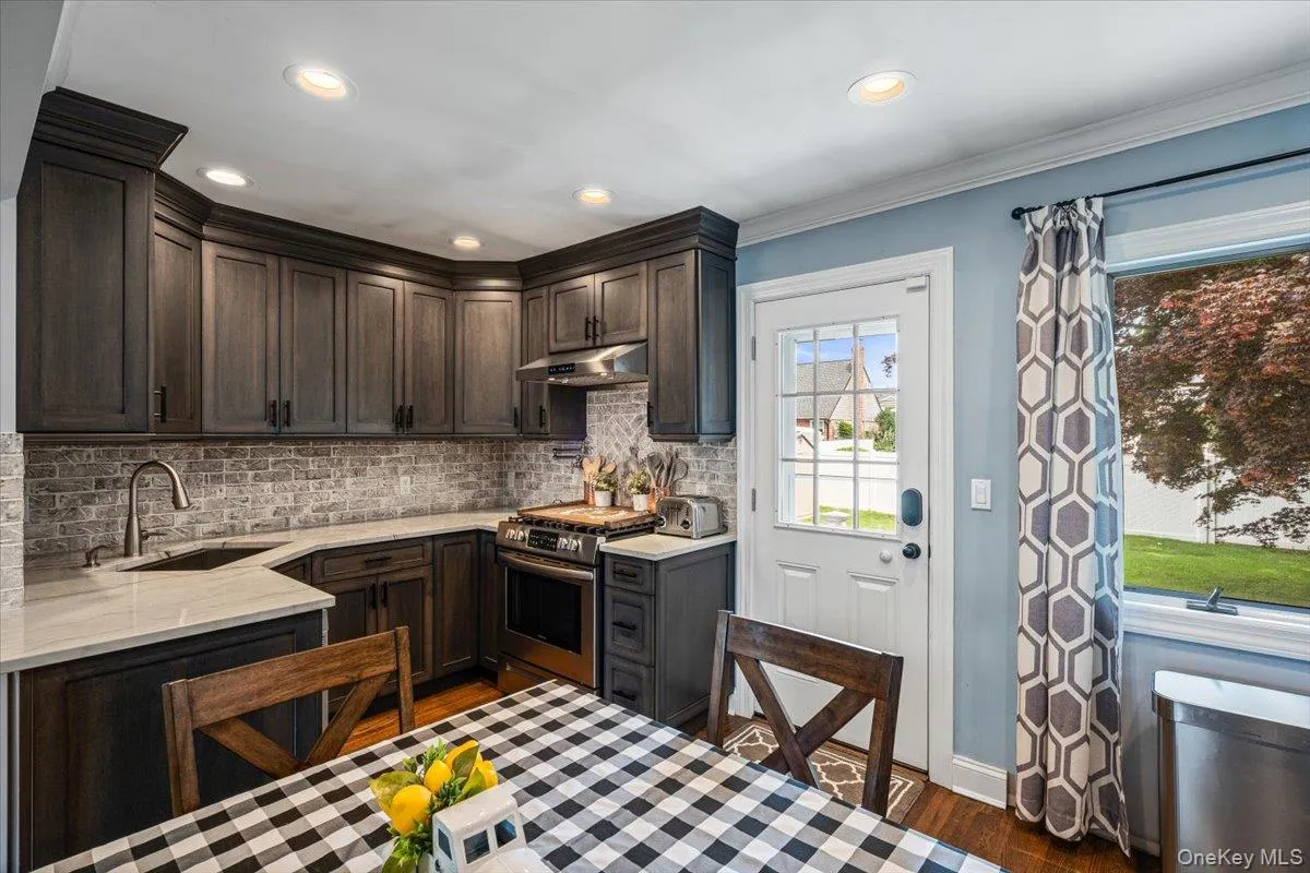 Dark wood-finish cabinetry with crown molding, light-toned countertops, a full-height tile backsplash, a stainless steel range with matching hood, and recessed lighting Dark wood-finish cabinetry with crown molding, light-toned countertops, a full-height tile backsplash, a stainless steel range with matching hood, and recessed lighting