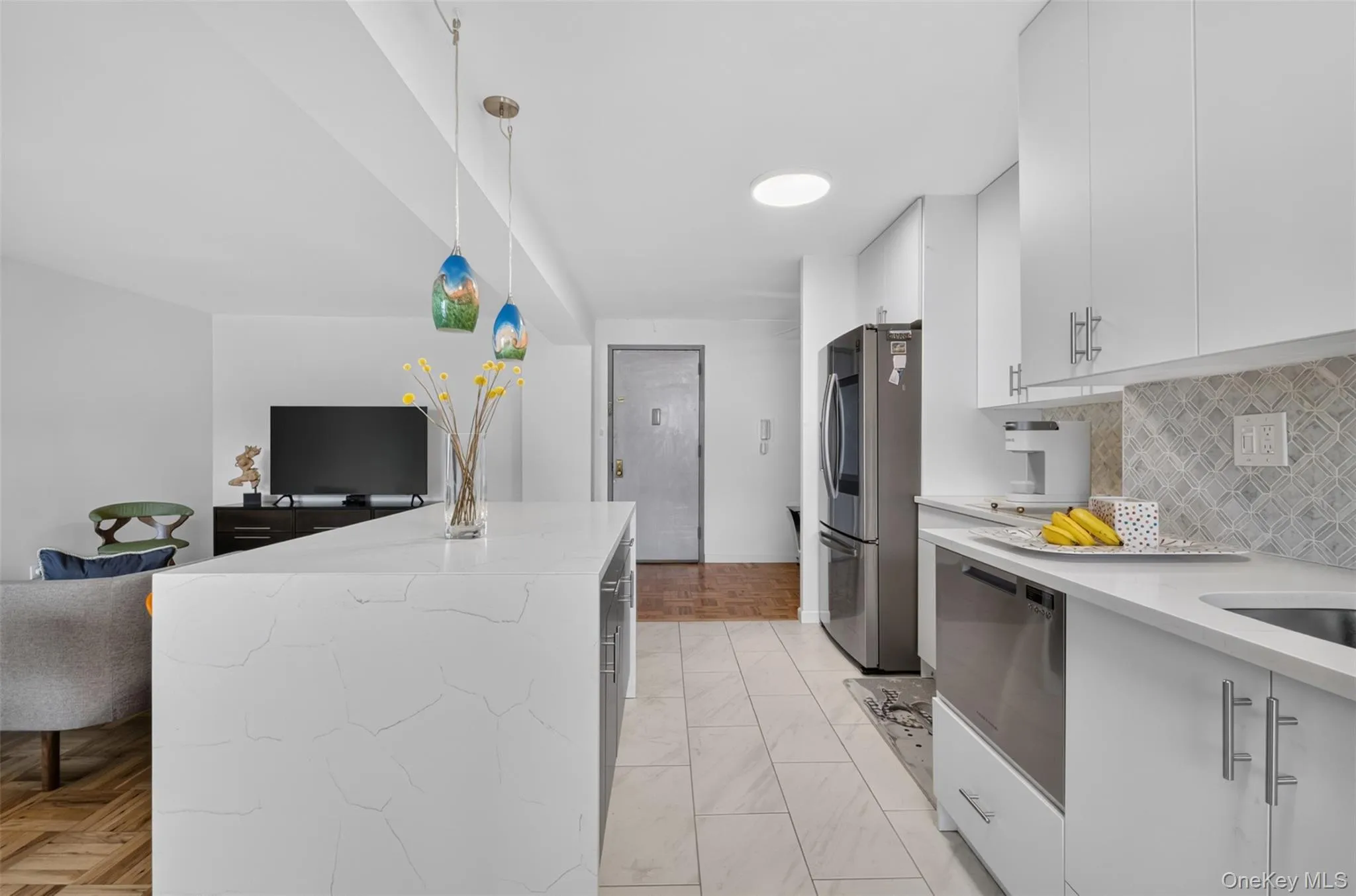 Integrated kitchen and living area featuring a large island with a light-toned countertop, white cabinetry with stainless steel hardware, and stainless steel appliances Integrated kitchen and living area featuring a large island with a light-toned countertop, white cabinetry with stainless steel hardware, and stainless steel appliances