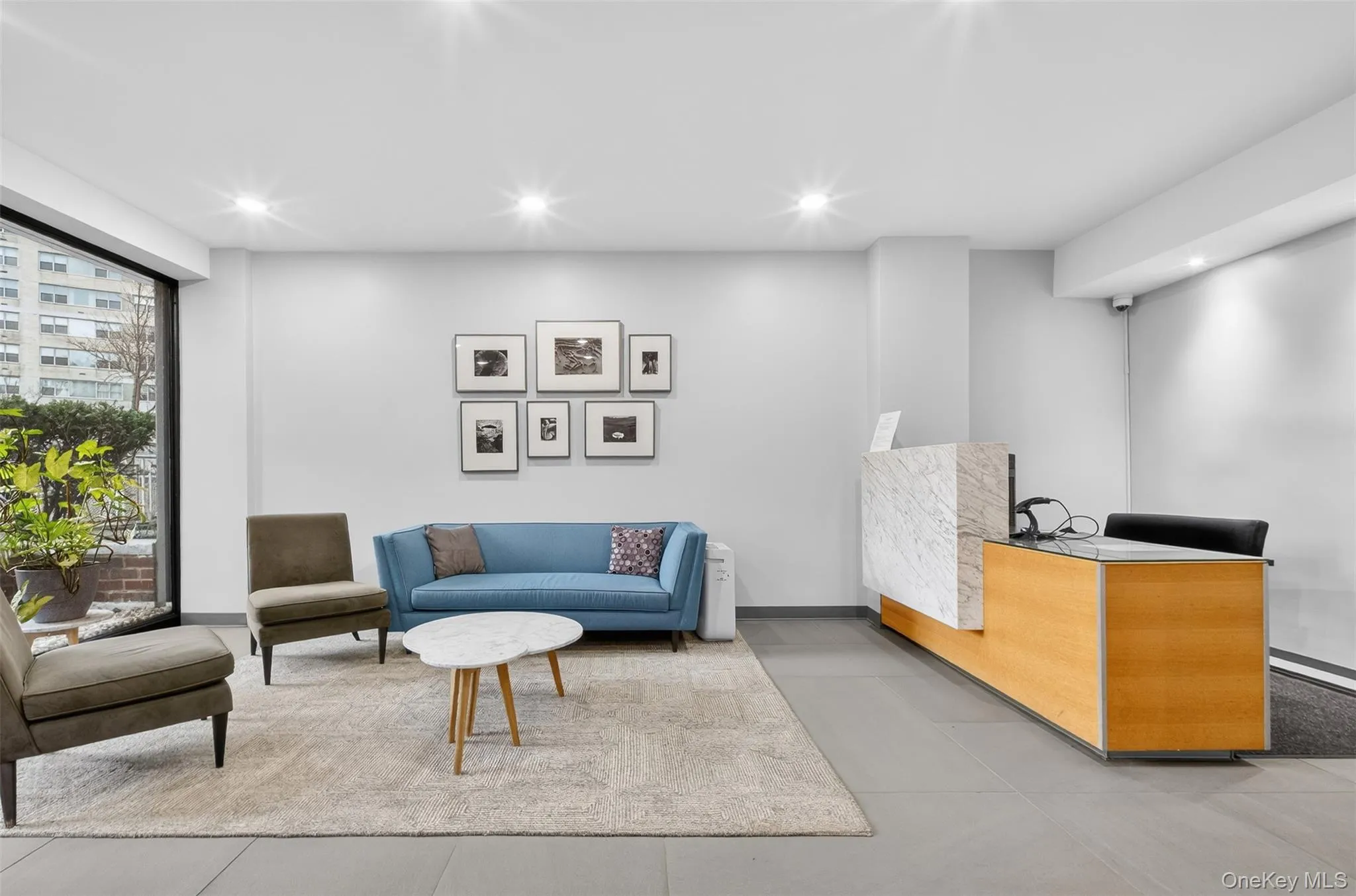 Expansive full-height windows, recessed ceiling lighting, a reception desk with a light wood finish and stone-look top, and gray tile flooring Expansive full-height windows, recessed ceiling lighting, a reception desk with a light wood finish and stone-look top, and gray tile flooring