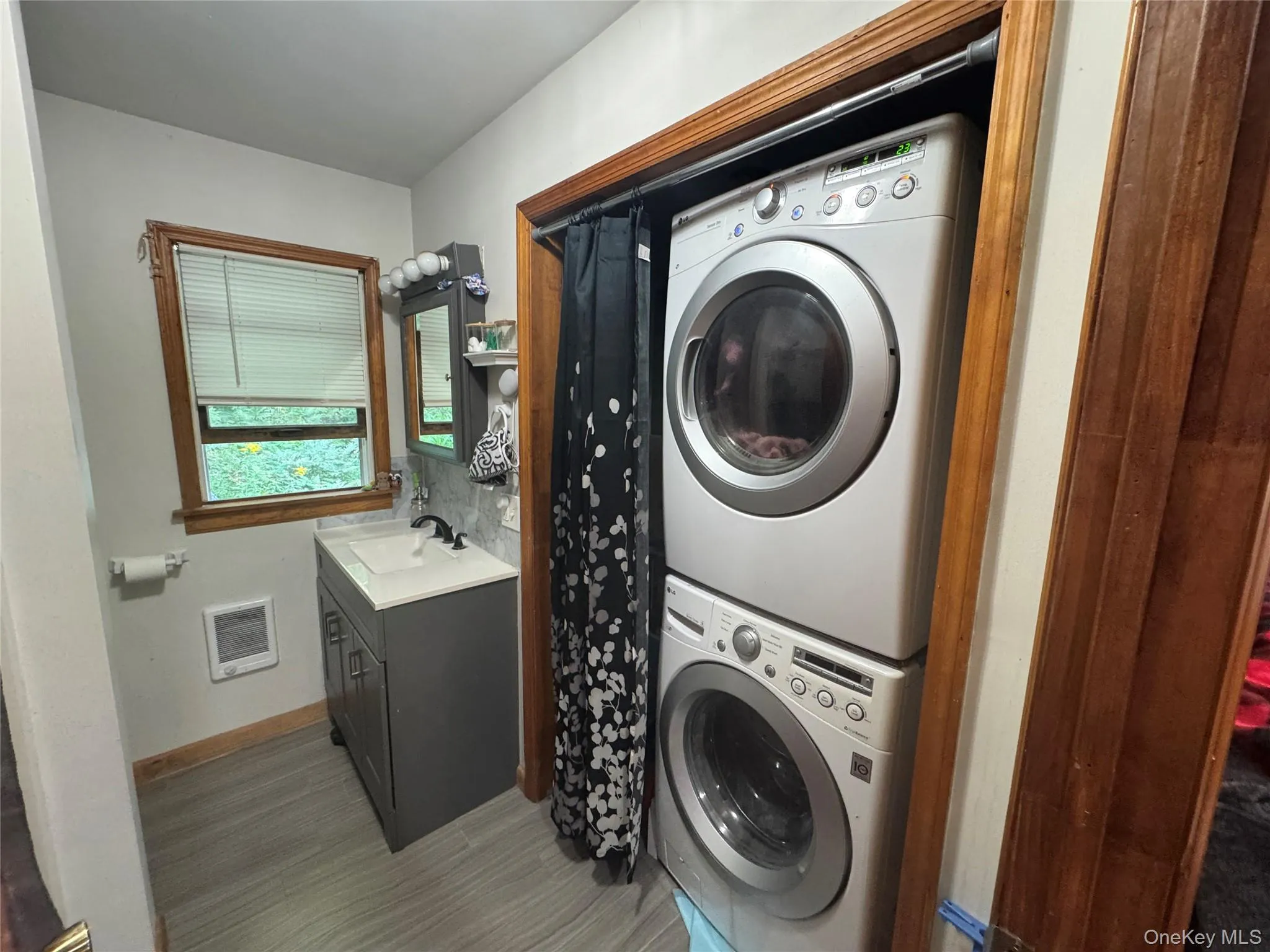 White stacked washer and dryer unit, modern vanity with white countertop and black faucet, light gray tile-look flooring, and a window with wood trim White stacked washer and dryer unit, modern vanity with white countertop and black faucet, light gray tile-look flooring, and a window with wood trim