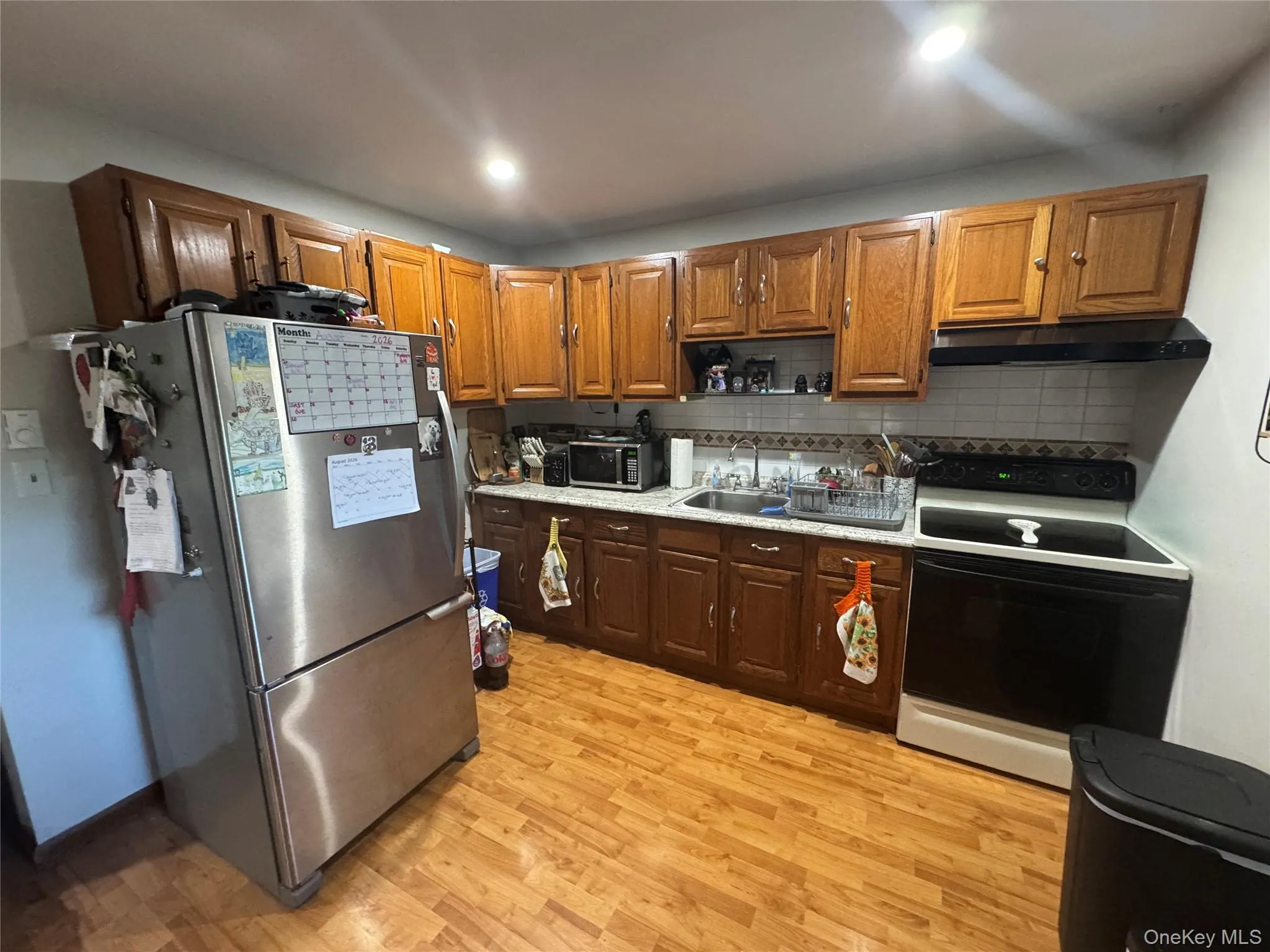 Wood-finish flooring, wood cabinetry, stainless steel refrigerator, white tile backsplash, and recessed lighting Wood-finish flooring, wood cabinetry, stainless steel refrigerator, white tile backsplash, and recessed lighting