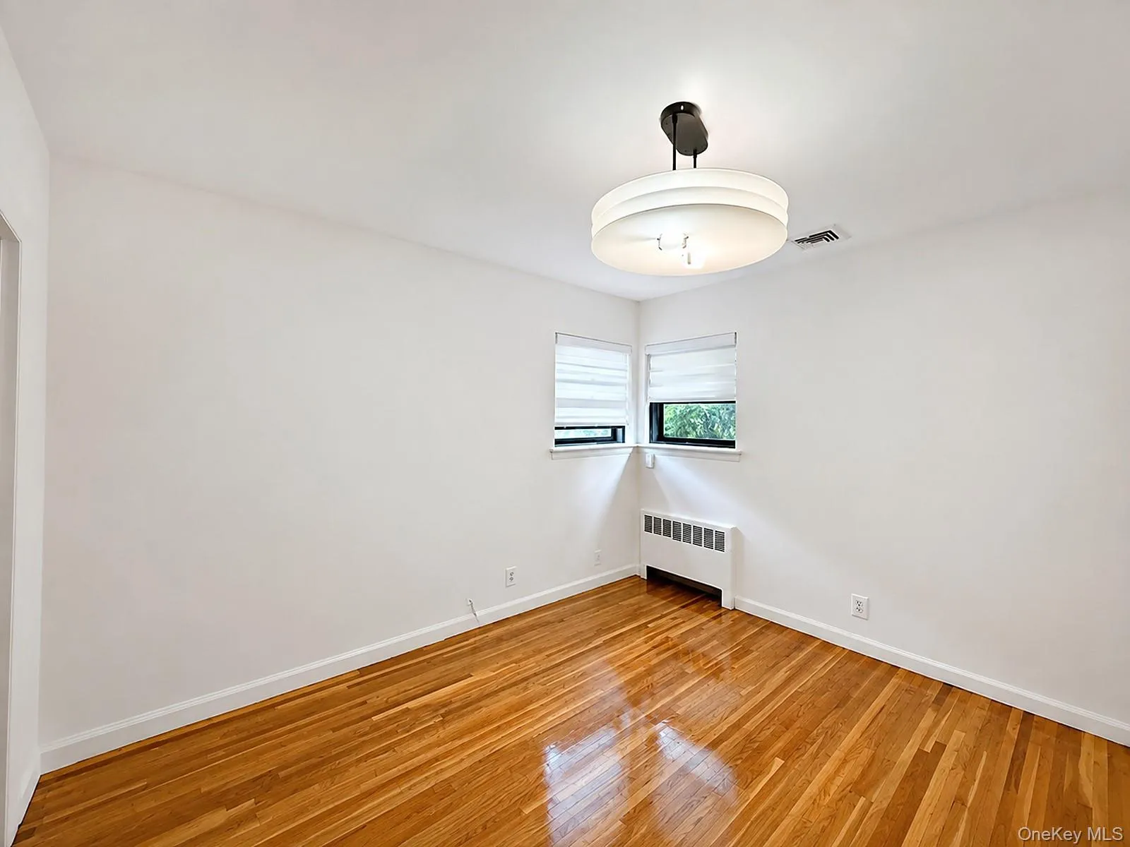 Hardwood flooring, window with roller shades, baseboard heating unit, and a flush-mount ceiling light fixture Hardwood flooring, window with roller shades, baseboard heating unit, and a flush-mount ceiling light fixture
