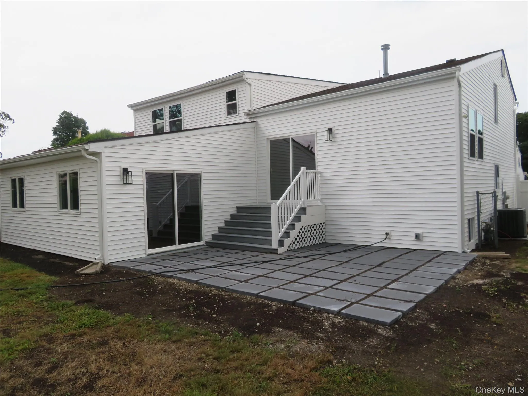 White siding exterior with multiple windows, a grey pavers patio, a multi-level deck with white railings, and sliding glass doors White siding exterior with multiple windows, a grey pavers patio, a multi-level deck with white railings, and sliding glass doors