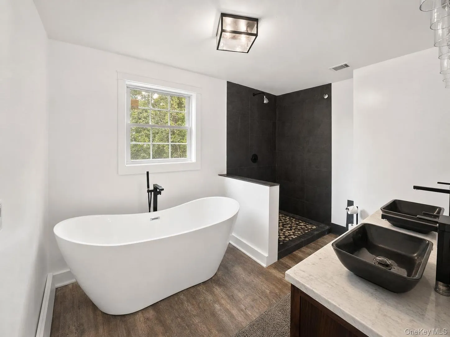 Freestanding soaking tub, wood-finish flooring, walk-in shower with dark tile surround and mosaic floor, vanity with two vessel sinks, and dark fixtures Freestanding soaking tub, wood-finish flooring, walk-in shower with dark tile surround and mosaic floor, vanity with two vessel sinks, and dark fixtures