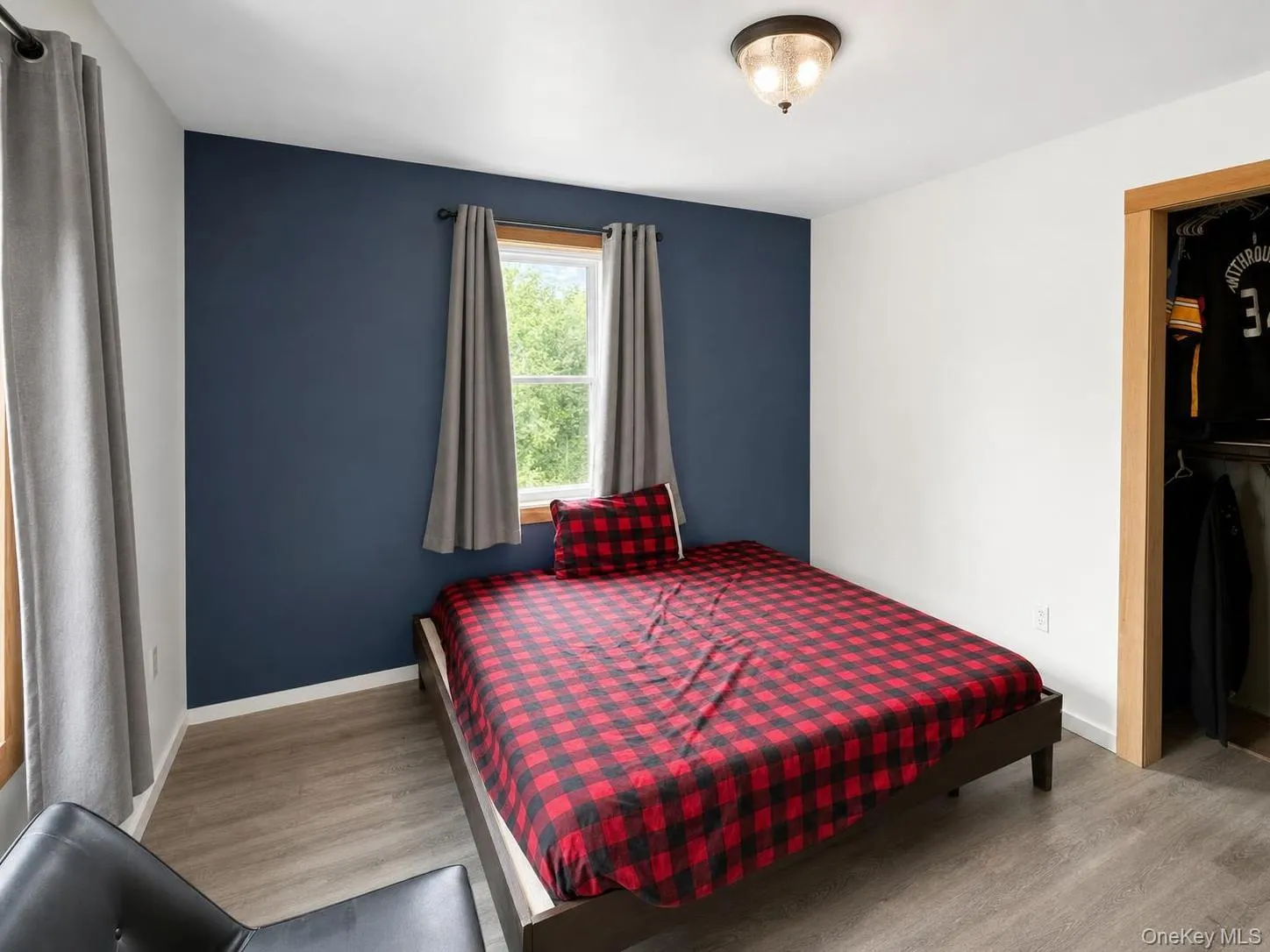 Wood-finish flooring, accent wall, window with natural wood trim, flush mount ceiling light, and a built-in closet with natural wood trim Wood-finish flooring, accent wall, window with natural wood trim, flush mount ceiling light, and a built-in closet with natural wood trim