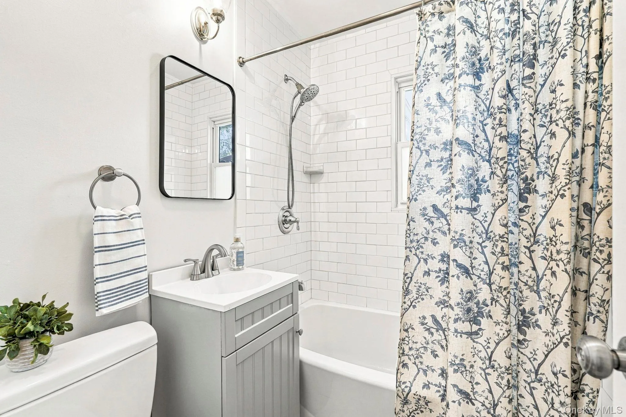 White subway tile shower surround, chrome shower fixtures, white bathtub, gray vanity with integrated sink, and wall-mounted mirror White subway tile shower surround, chrome shower fixtures, white bathtub, gray vanity with integrated sink, and wall-mounted mirror