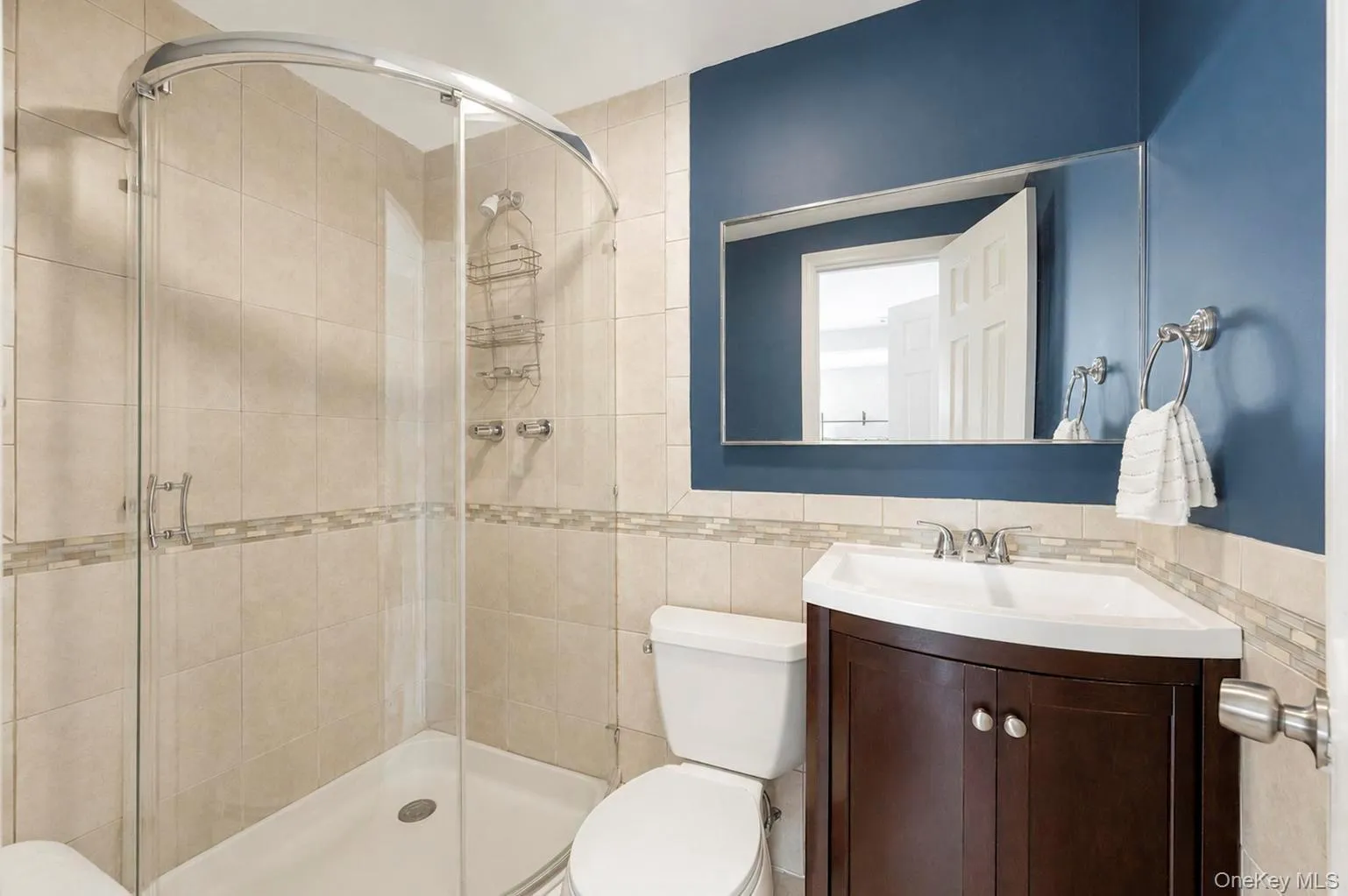 Corner shower with clear glass enclosure, ceramic wall tiling with accent banding, corner vanity with dark wood-finish cabinetry, white basin, and a wall-mounted mirror Corner shower with clear glass enclosure, ceramic wall tiling with accent banding, corner vanity with dark wood-finish cabinetry, white basin, and a wall-mounted mirror