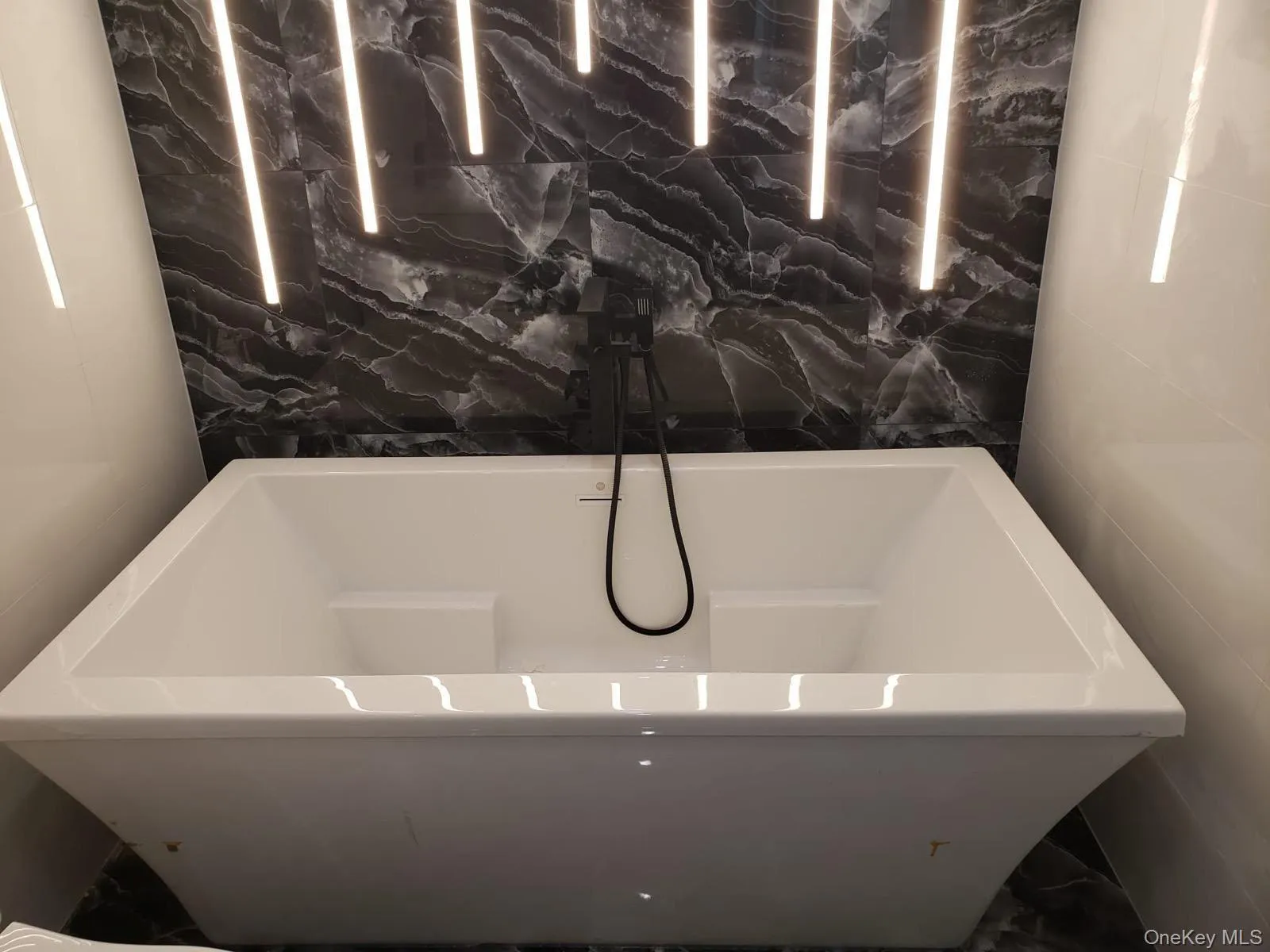 Freestanding rectangular soaking tub with a matte black wall-mounted faucet, set against a dark veined accent wall with integrated linear lighting Freestanding rectangular soaking tub with a matte black wall-mounted faucet, set against a dark veined accent wall with integrated linear lighting