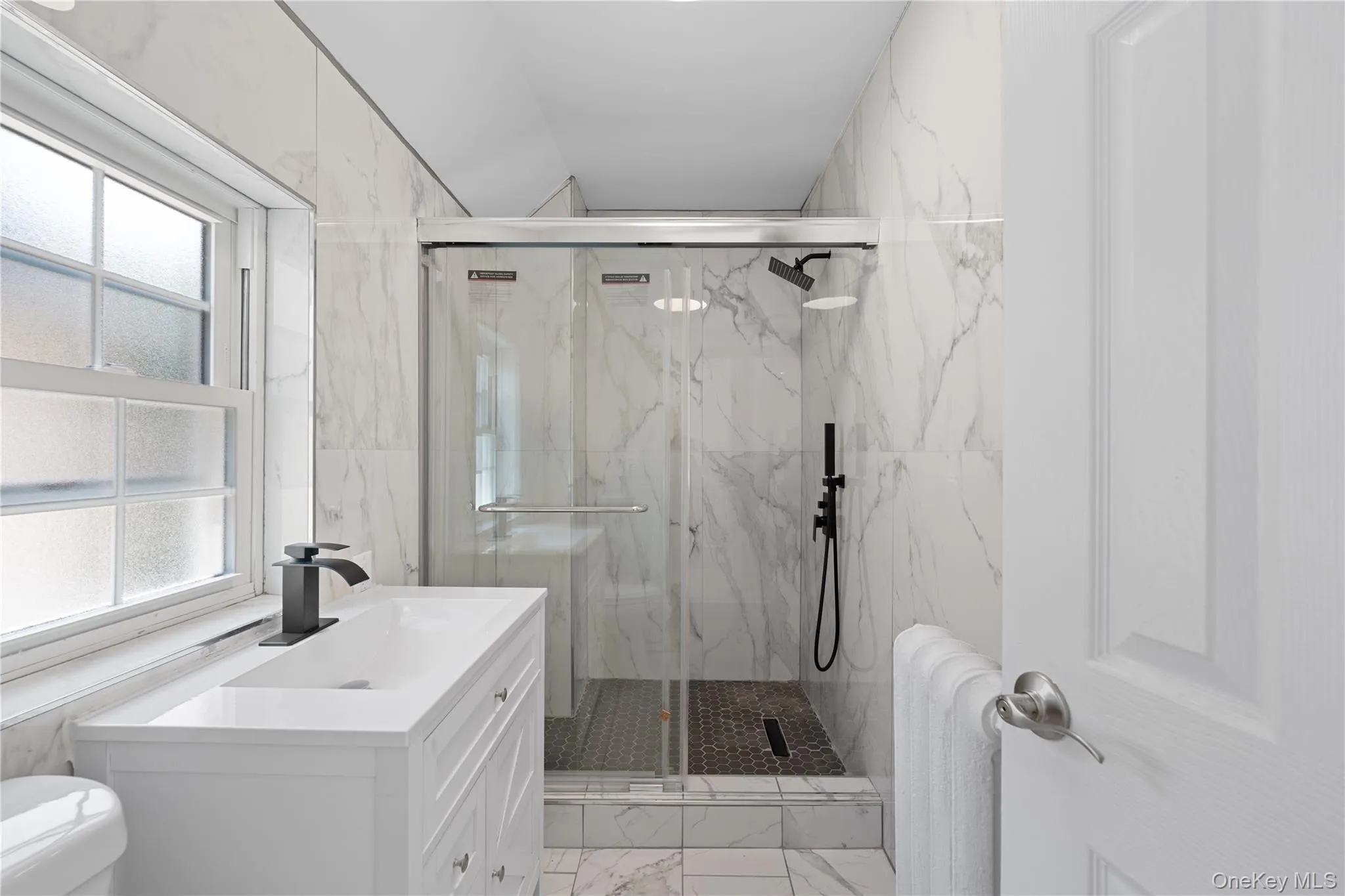 white vanity with integrated rectangular sink and matte black faucet, glass-enclosed shower, matte black shower fixtures, and white marble-look wall tiling white vanity with integrated rectangular sink and matte black faucet, glass-enclosed shower, matte black shower fixtures, and white marble-look wall tiling