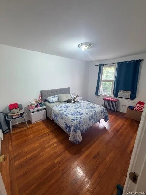 Spacious bedroom featuring hardwood flooring, large window with natural light, ceiling-mounted light fixture, and radiator heating Spacious bedroom featuring hardwood flooring, large window with natural light, ceiling-mounted light fixture, and radiator heating