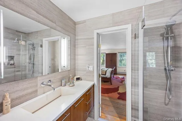 Bathroom featuring a frameless glass-enclosed shower, wall-mounted shower fixture, an integrated rectangular sink, wood-finish cabinetry, and a large vanity mirror Bathroom featuring a frameless glass-enclosed shower, wall-mounted shower fixture, an integrated rectangular sink, wood-finish cabinetry, and a large vanity mirror