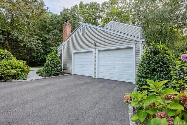 Side entry two-car garage with panel doors, asphalt driveway, brick chimney, and mature perimeter landscaping Side entry two-car garage with panel doors, asphalt driveway, brick chimney, and mature perimeter landscaping