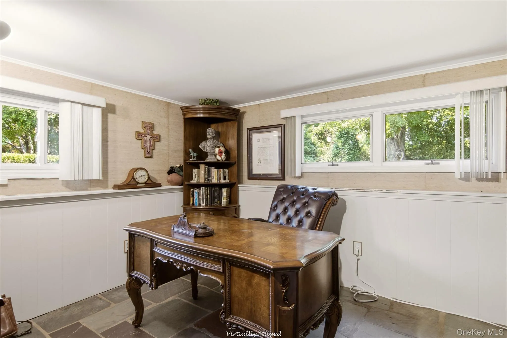 Sophisticated home office featuring a wood-finish desk with carved legs, built-in shelving with display space, large multi-pane windows with natural light, neutral two-tone wall treatment with wainscoting, and slate-finish flooring Sophisticated home office featuring a wood-finish desk with carved legs, built-in shelving with display space, large multi-pane windows with natural light, neutral two-tone wall treatment with wainscoting, and slate-finish flooring