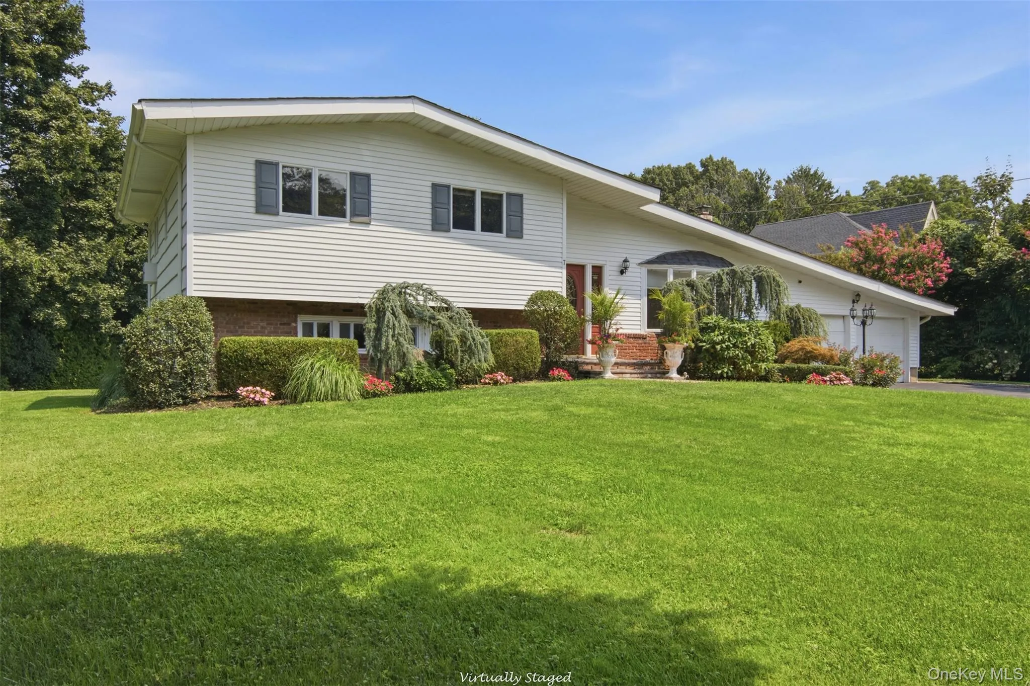 White siding exterior with a brick lower level, featuring a two-car garage, mature landscaping, and an expansive lawn White siding exterior with a brick lower level, featuring a two-car garage, mature landscaping, and an expansive lawn