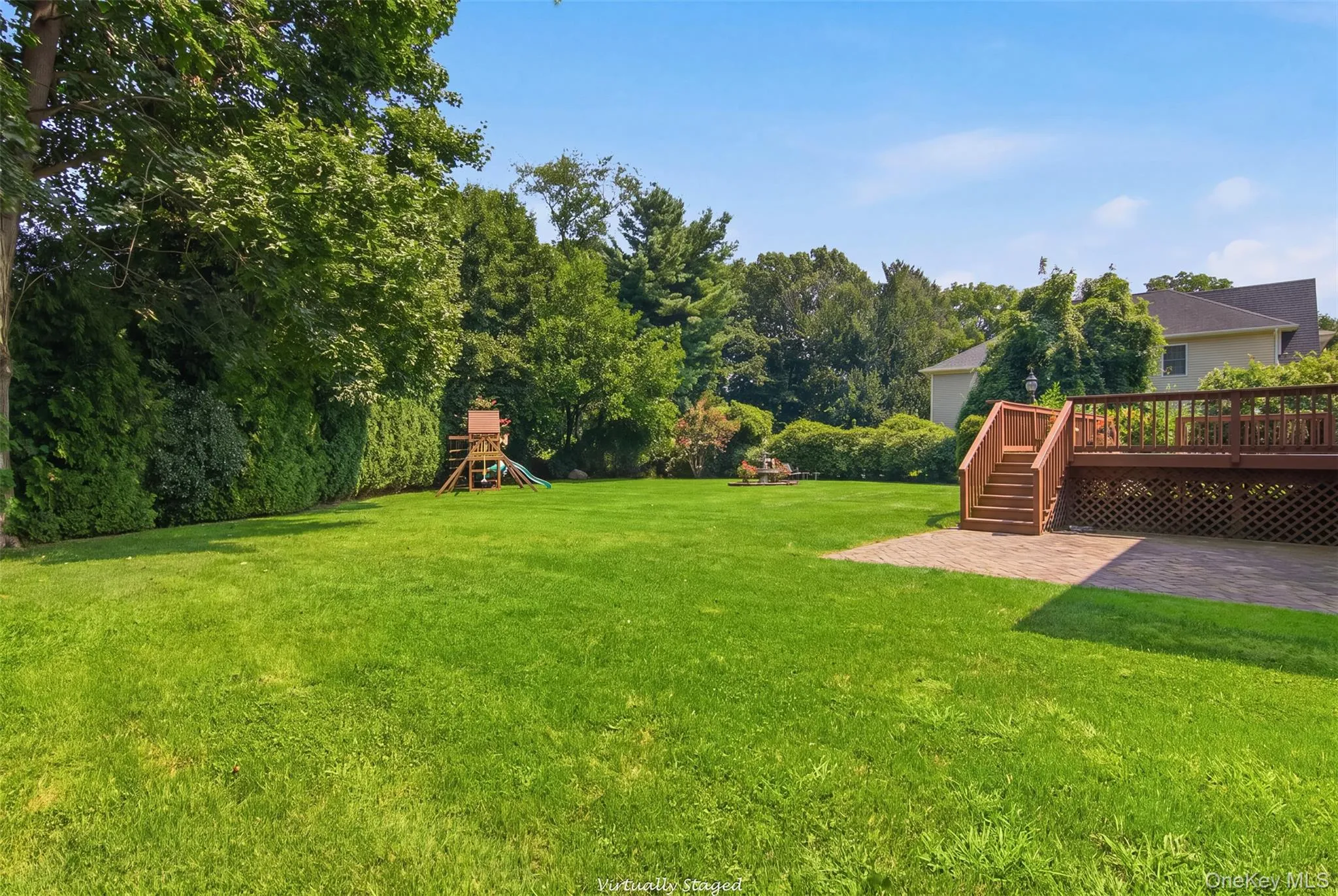 Expansive manicured lawn with mature trees, elevated wood deck with stairs, and a paver patio Expansive manicured lawn with mature trees, elevated wood deck with stairs, and a paver patio