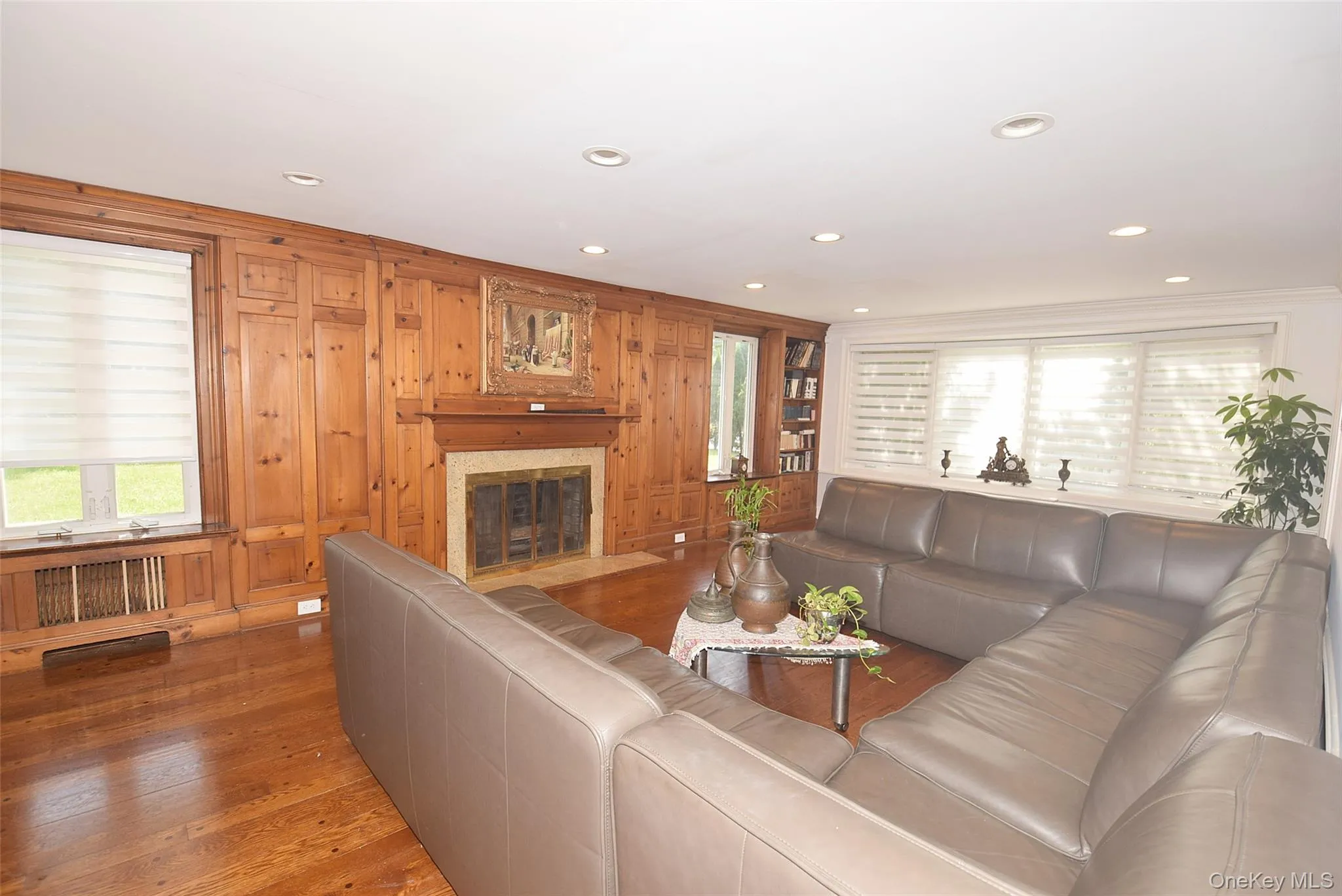 Wood-paneled wall with a central fireplace and mantel, recessed ceiling lighting, wood-finish flooring, built-in shelving, and multiple windows Wood-paneled wall with a central fireplace and mantel, recessed ceiling lighting, wood-finish flooring, built-in shelving, and multiple windows