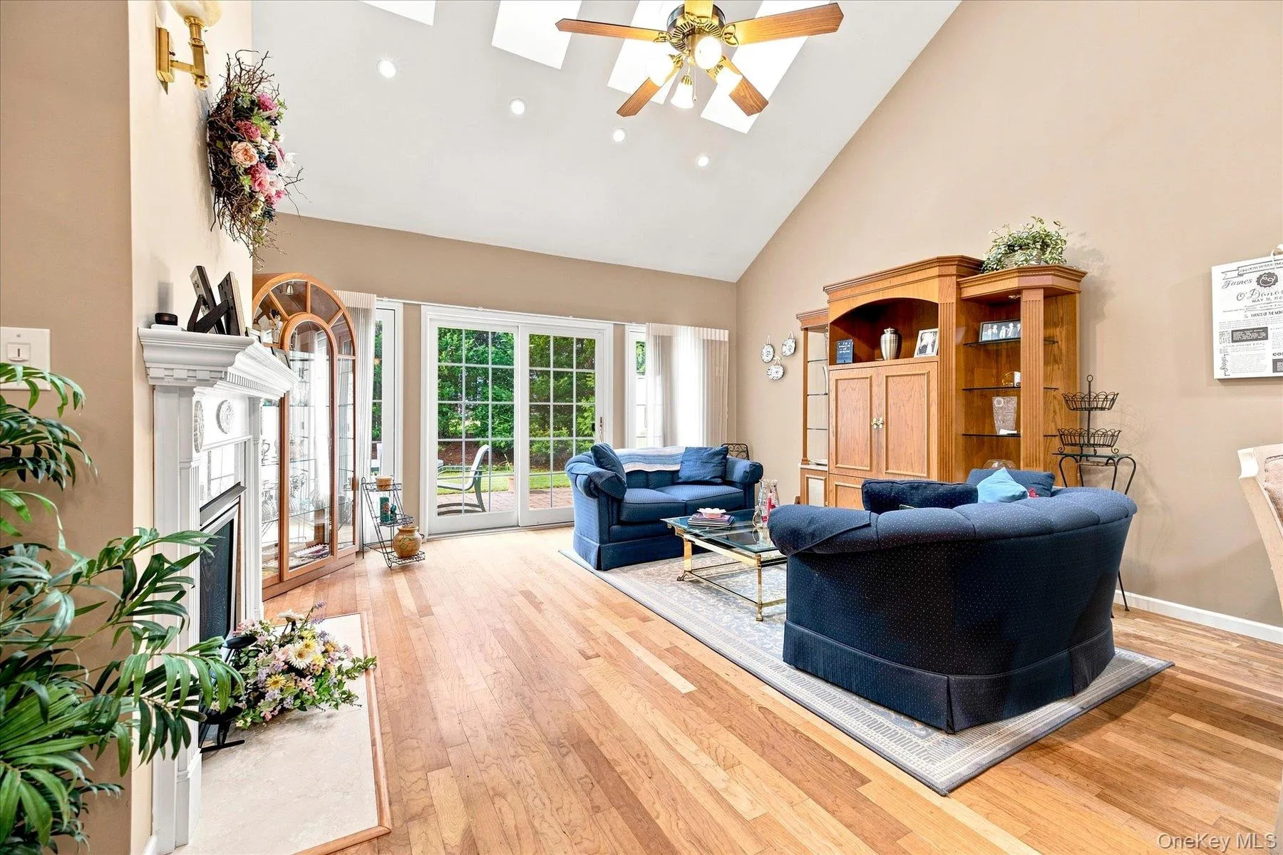 Vaulted ceiling with skylights and ceiling fan, wood-finish flooring, white fireplace with decorative mantel, and glass paneled patio doors Vaulted ceiling with skylights and ceiling fan, wood-finish flooring, white fireplace with decorative mantel, and glass paneled patio doors