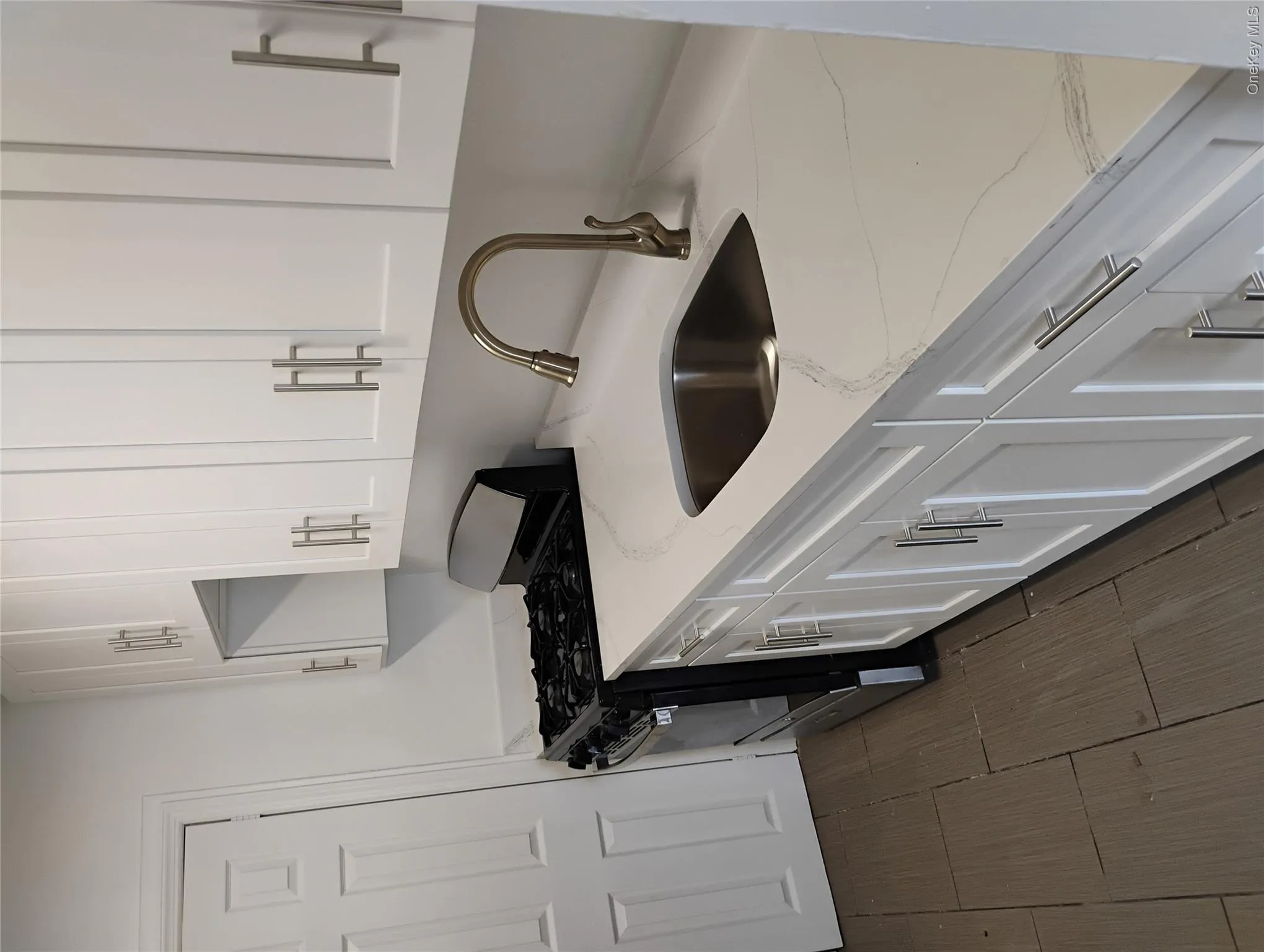 White shaker-style cabinetry with brushed nickel hardware, stainless steel undermount sink, gooseneck faucet, white countertop with veining, and dark-finish tile flooring White shaker-style cabinetry with brushed nickel hardware, stainless steel undermount sink, gooseneck faucet, white countertop with veining, and dark-finish tile flooring