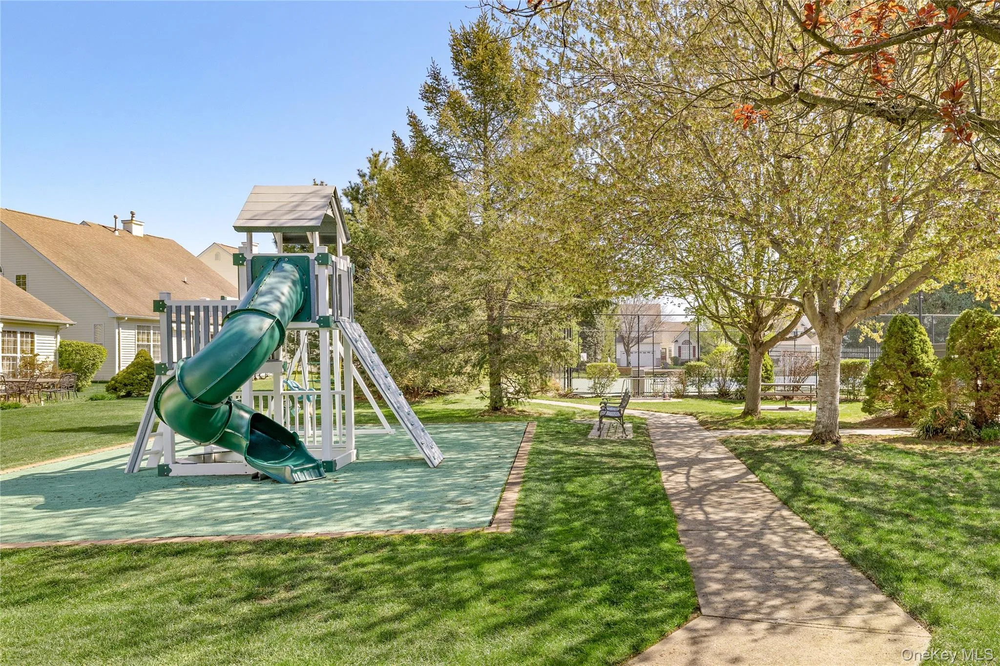 Outdoor recreational area featuring a playground structure with a green slide, a concrete pathway, a park bench, and mature trees Outdoor recreational area featuring a playground structure with a green slide, a concrete pathway, a park bench, and mature trees