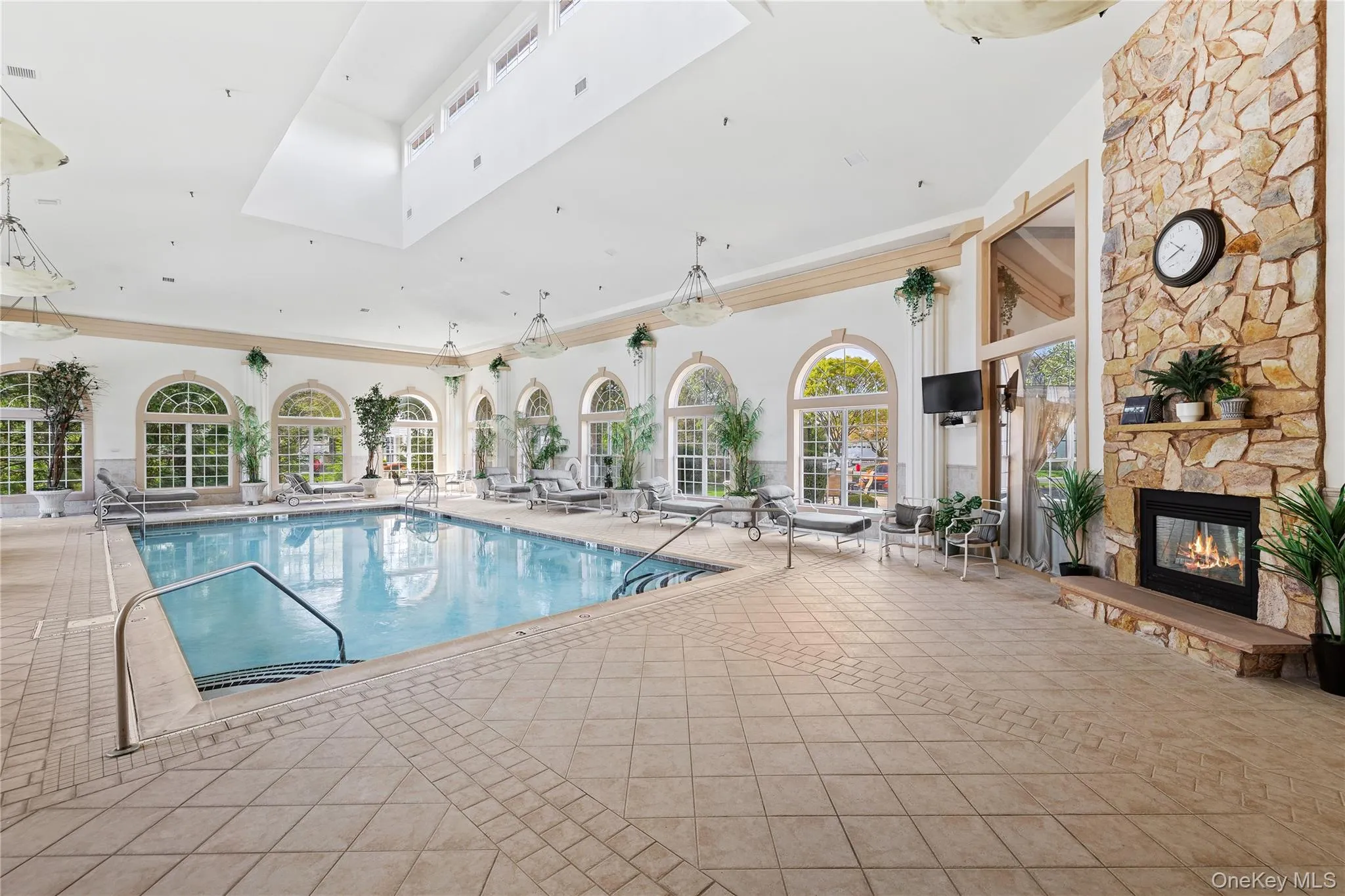 Indoor swimming pool with integrated spa, large arched windows, stone-clad fireplace with hearth, and tiled flooring Indoor swimming pool with integrated spa, large arched windows, stone-clad fireplace with hearth, and tiled flooring