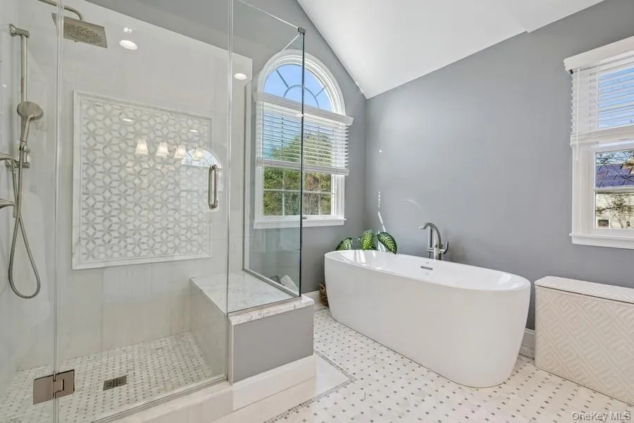 Spa-inspired primary bathroom featuring a freestanding soaking tub, walk-in shower with geometric tile surround and frameless glass enclosure, mosaic tile flooring, dual windows with arched transom, and polished nickel fixtures throughout Spa-inspired primary bathroom featuring a freestanding soaking tub, walk-in shower with geometric tile surround and frameless glass enclosure, mosaic tile flooring, dual windows with arched transom, and polished nickel fixtures throughout