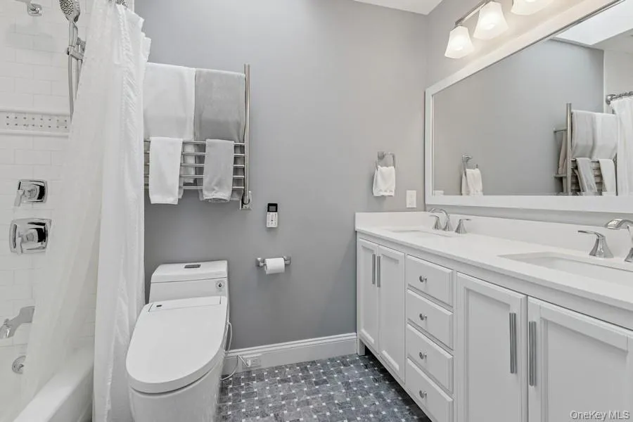 Spa-inspired bathroom featuring a dual-sink vanity with brushed nickel hardware, white cabinetry with ample storage, quartz countertops, and a large frameless mirror with integrated lighting Spa-inspired bathroom featuring a dual-sink vanity with brushed nickel hardware, white cabinetry with ample storage, quartz countertops, and a large frameless mirror with integrated lighting