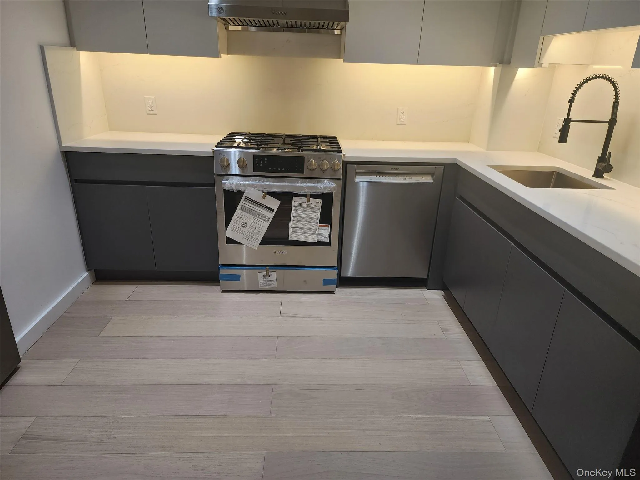 Kitchen featuring modern gray cabinetry, light-toned countertops, stainless steel appliances, and wood-finish flooring Kitchen featuring modern gray cabinetry, light-toned countertops, stainless steel appliances, and wood-finish flooring