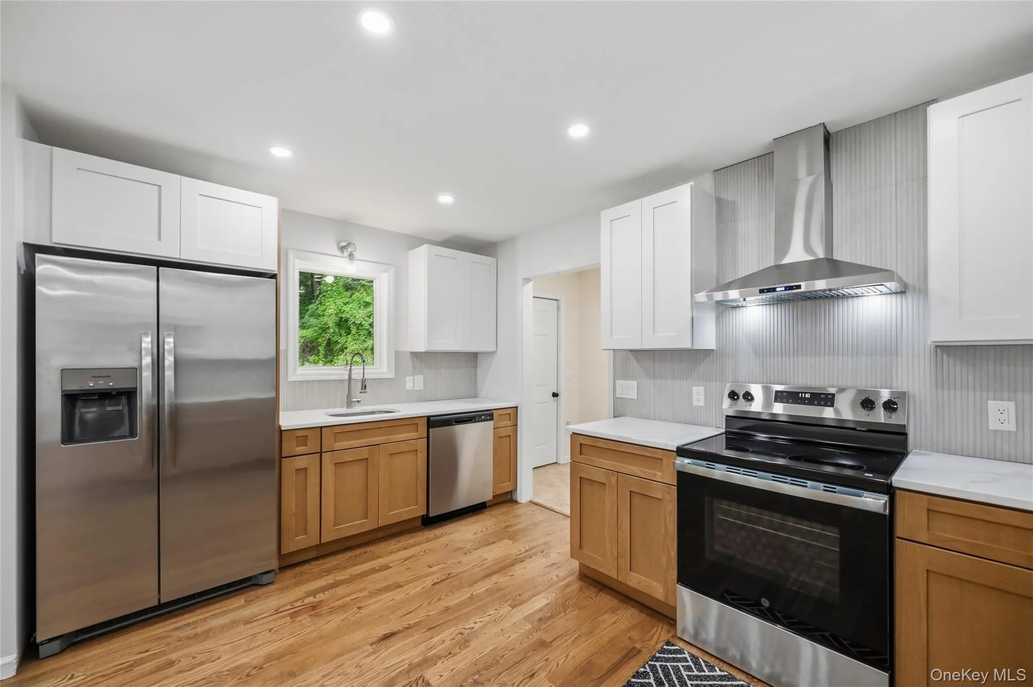 Kitchen featuring wood-finish flooring, stainless steel appliances, light wood-tone base cabinetry, white upper cabinetry, and light-colored countertops Kitchen featuring wood-finish flooring, stainless steel appliances, light wood-tone base cabinetry, white upper cabinetry, and light-colored countertops