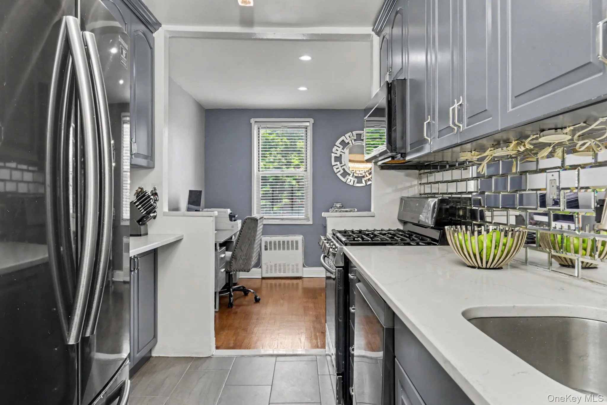 Grey cabinetry with brushed nickel hardware, stainless steel refrigerator, white countertops, mirrored backsplash, and stainless steel sink Grey cabinetry with brushed nickel hardware, stainless steel refrigerator, white countertops, mirrored backsplash, and stainless steel sink
